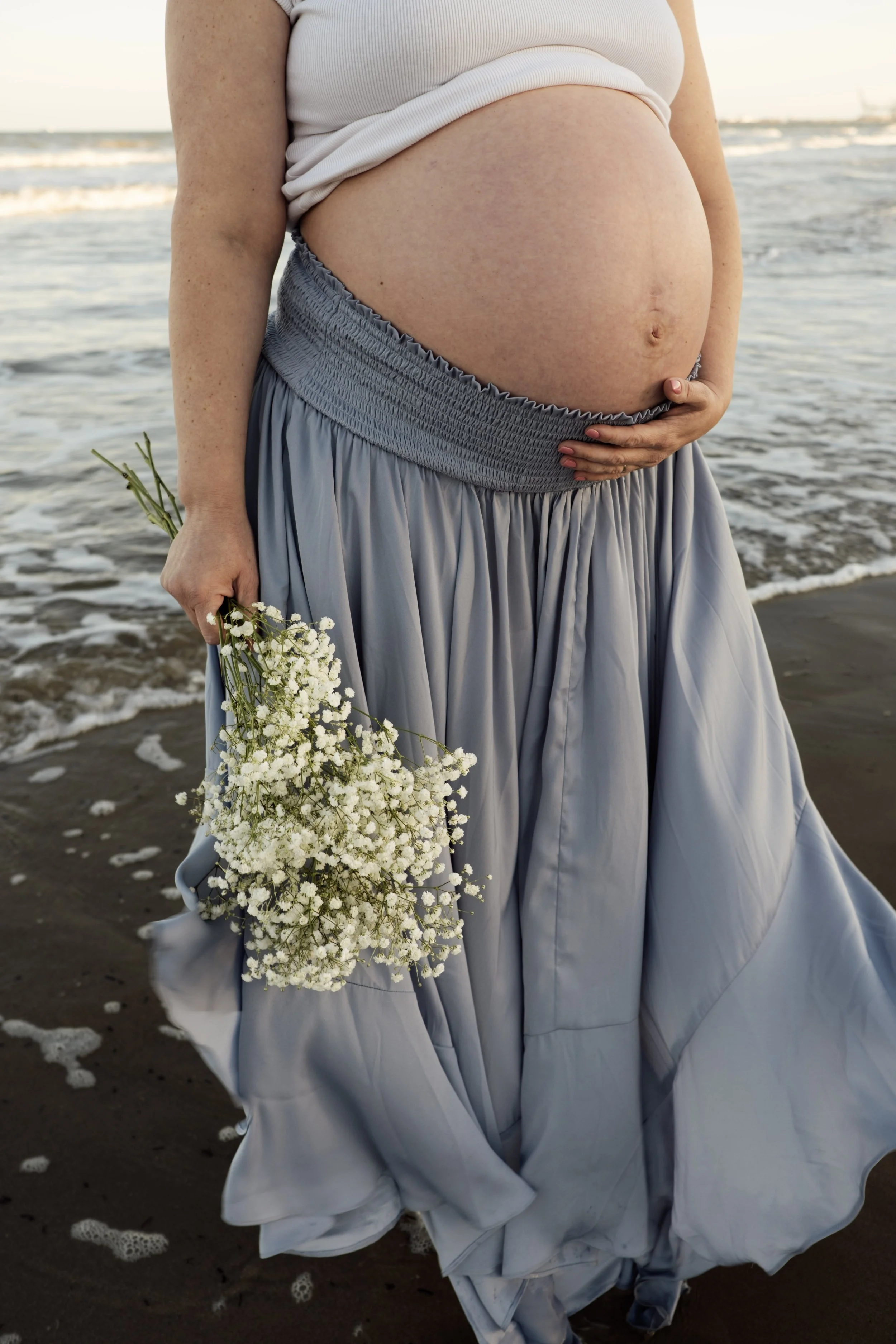 Pregnant woman in a flowing blue dress holding baby's breath flowers by the ocean