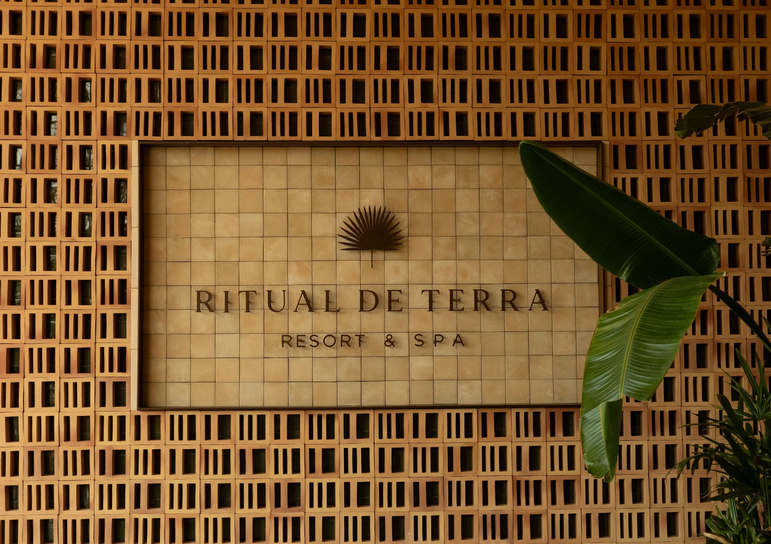 A wall with a decorative sign for Ritual De Terra Resort & Spa, featuring a palm leaf logo, surrounded by a lattice-style brick wall and green tropical plants.