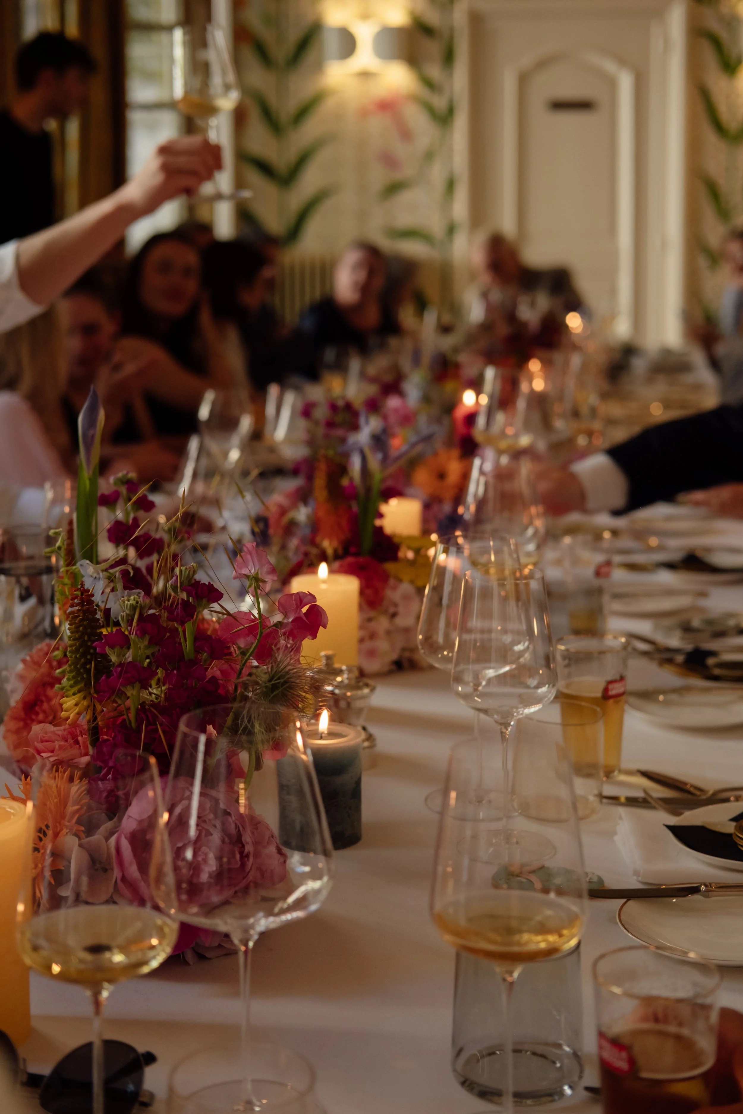 A long dinner table decorated with colorful flowers and lit candles, set for a formal event, with people sitting and a person raising a glass for a toast.