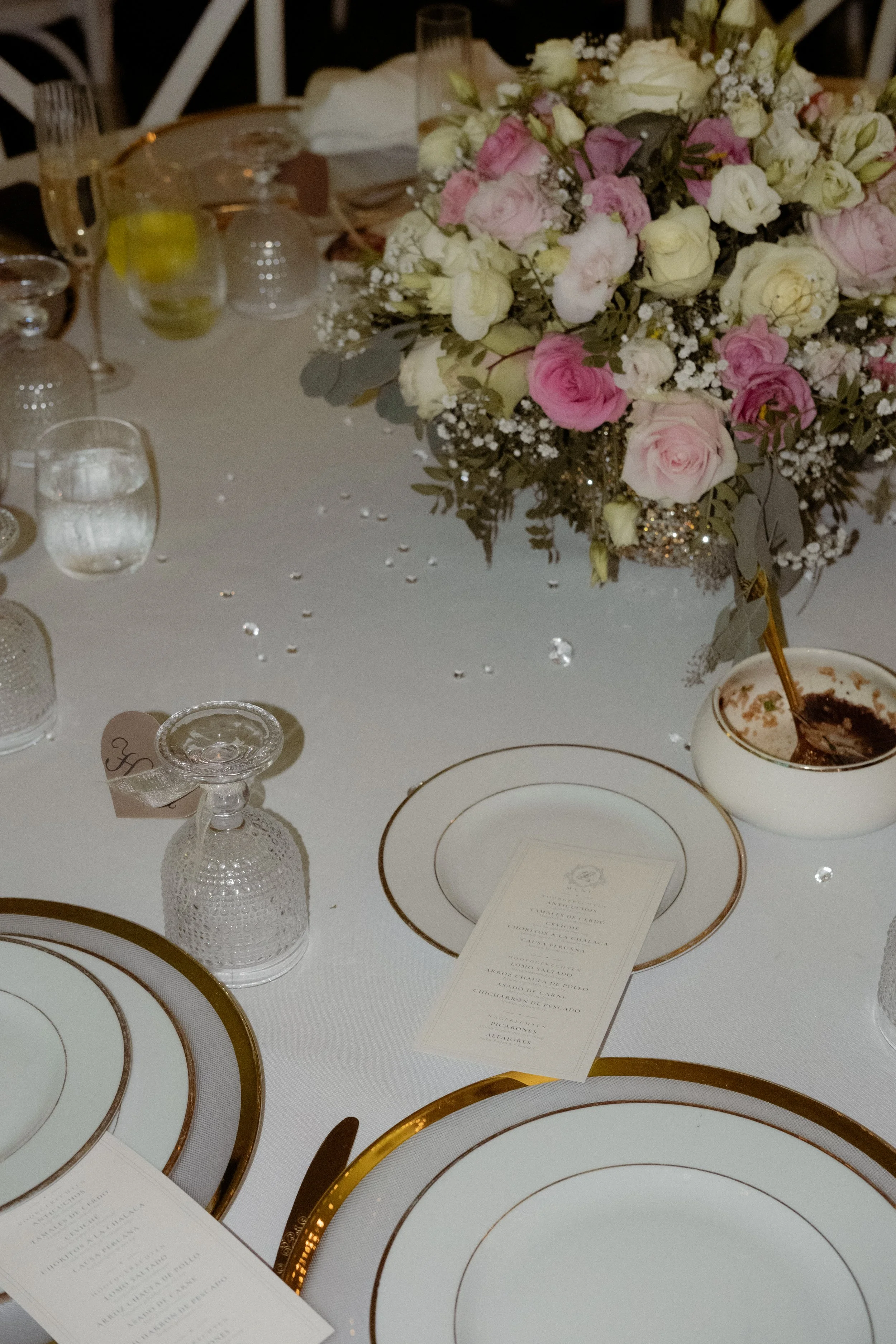 A table setting with white plates with gold rims, clear glassware, and a large floral centerpiece with white and pink roses, baby's breath, and greenery. There are also printed menus on the table.
