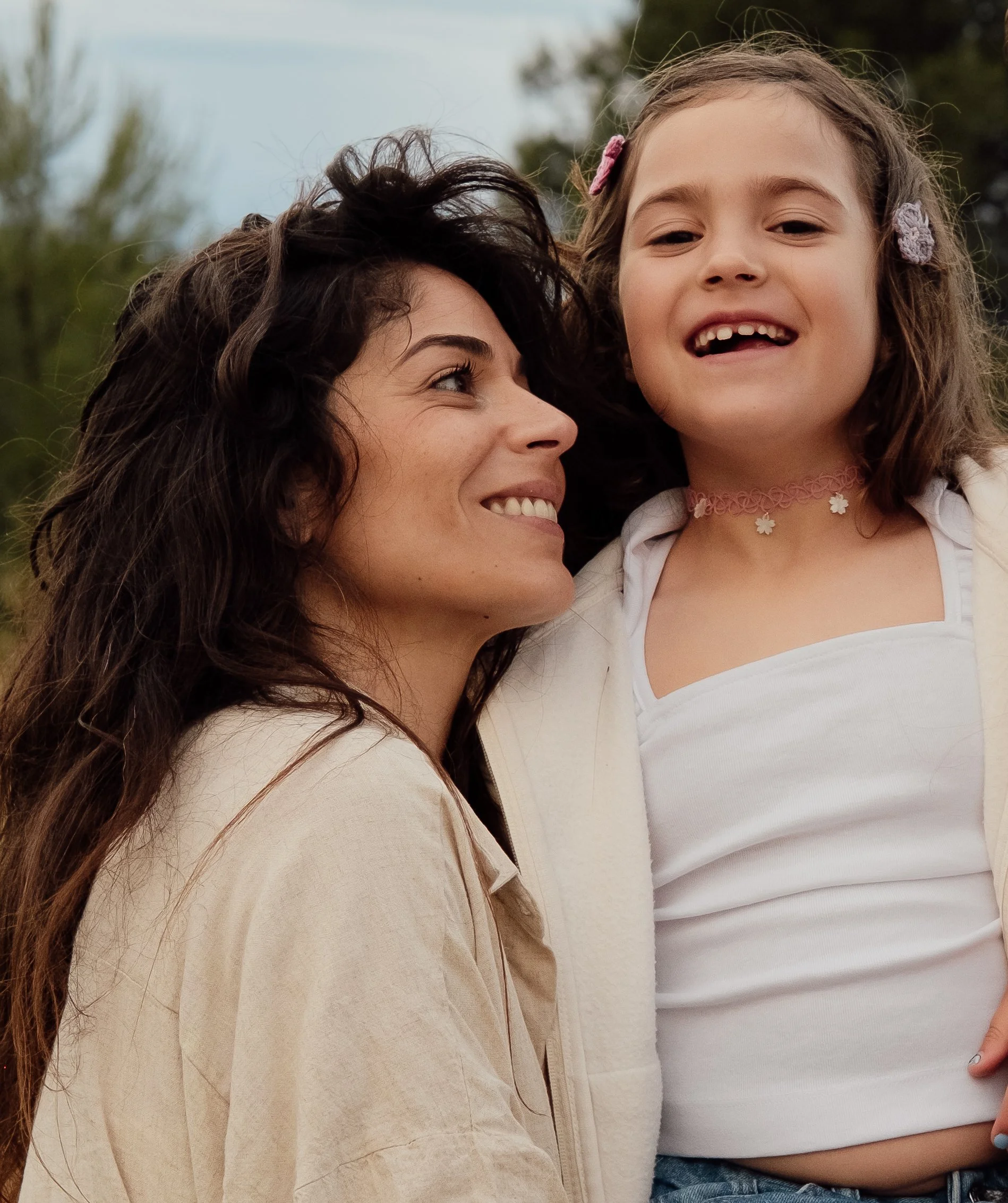 Two smiling women, one with long brown hair and the other with curly hair, are standing outdoors next to each other. The woman with long hair is looking up at the woman with curly hair, who has a big smile and missing front teeth. The scene appears t