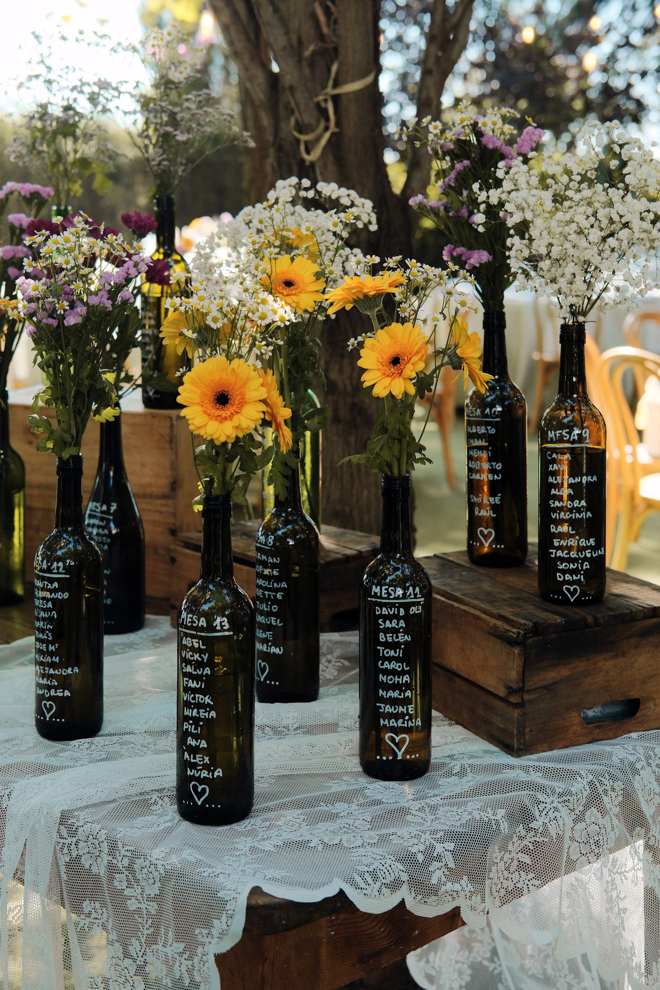 Glass bottles with handwritten names and messages used as vases holding colorful flowers on a lace-covered table, outdoors.