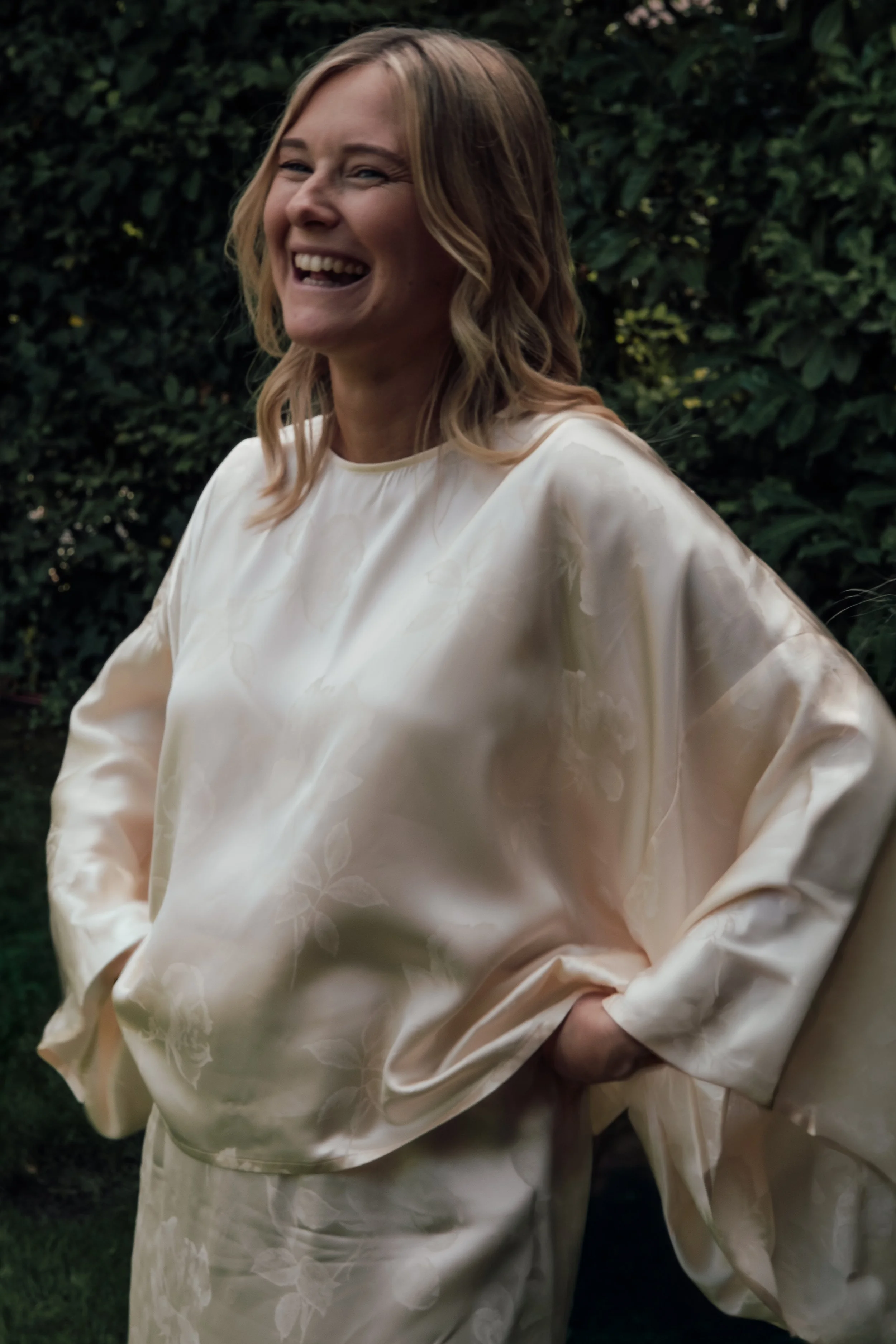 A woman smiling and laughing outdoors, wearing a cream-colored satin dress with a floral pattern, standing in front of greenery.