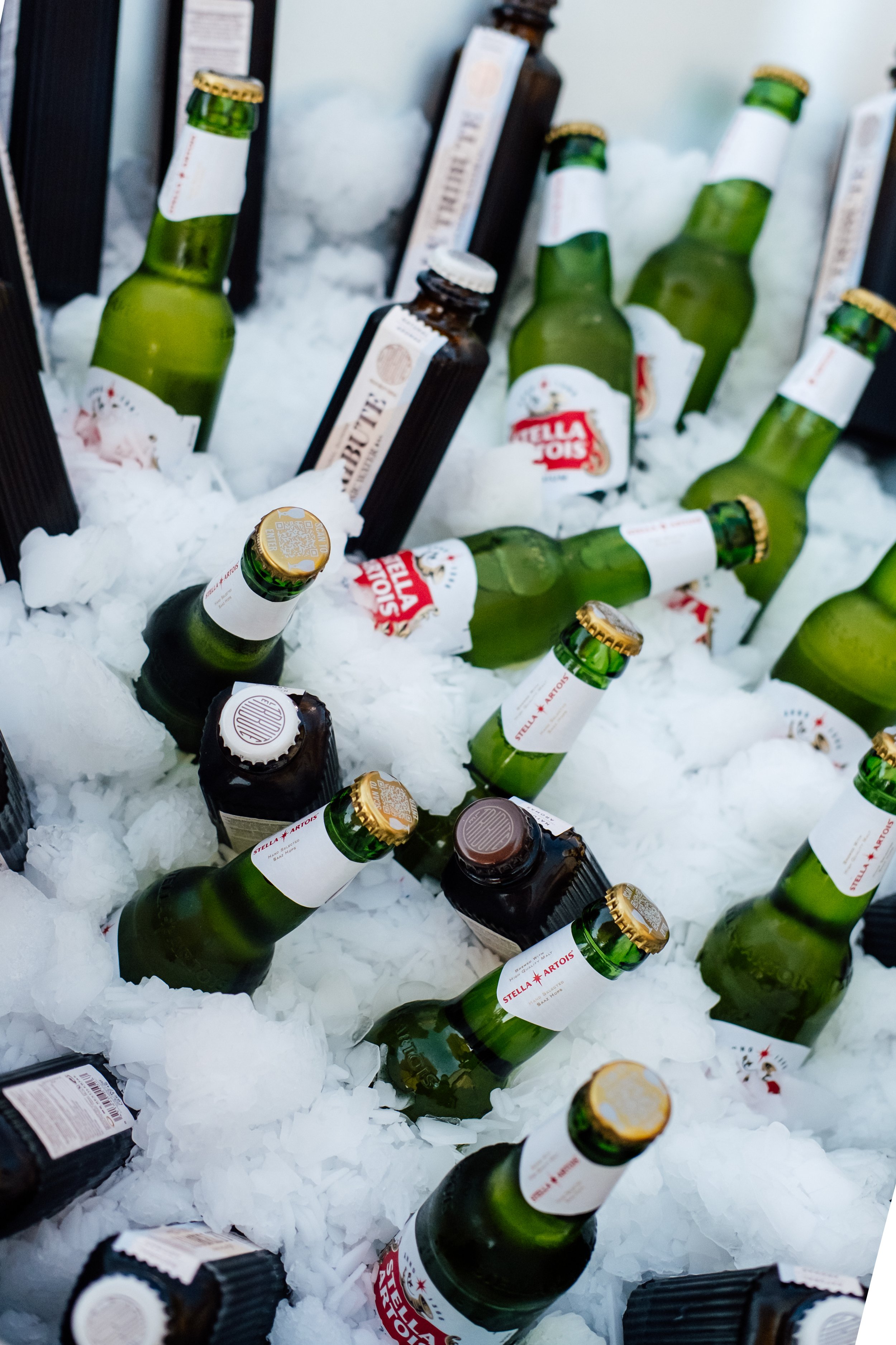 Green glass bottles of Stella Artois beer with white labels and gold caps, placed on a bed of ice in a cooler, with some dark brown bottles visible in the background.