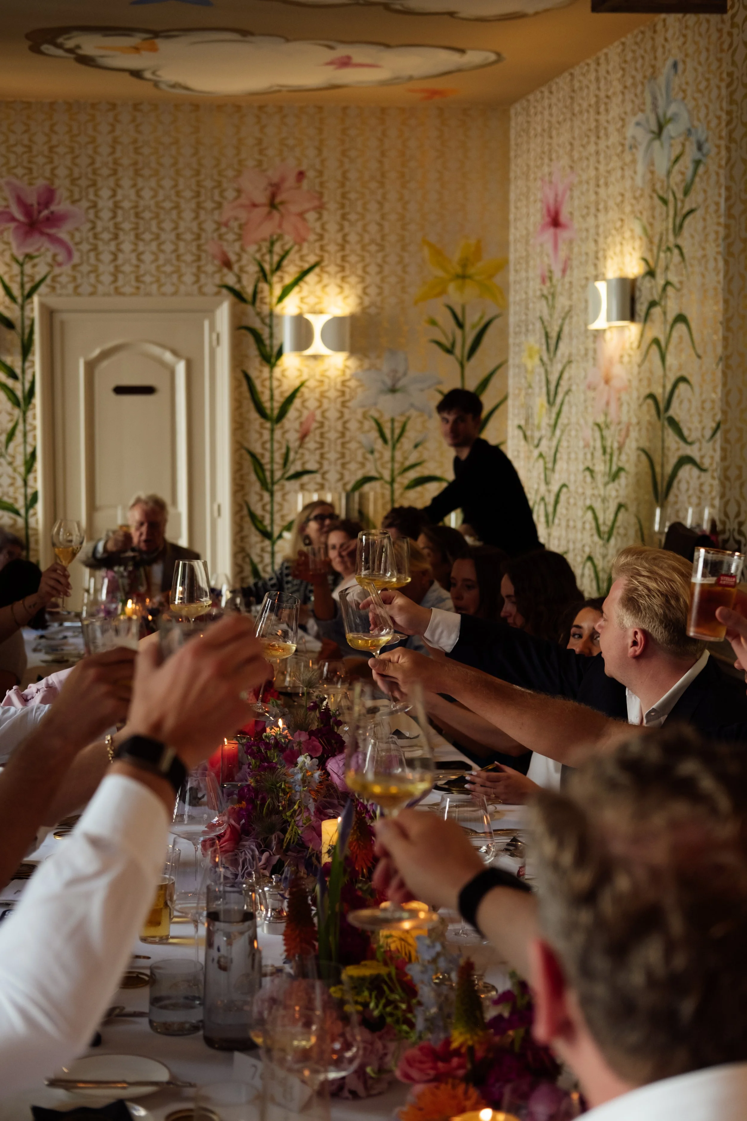 People gathered around a long dining table raising glasses for a toast at a celebration or formal dinner, with floral centerpieces and decorated walls.