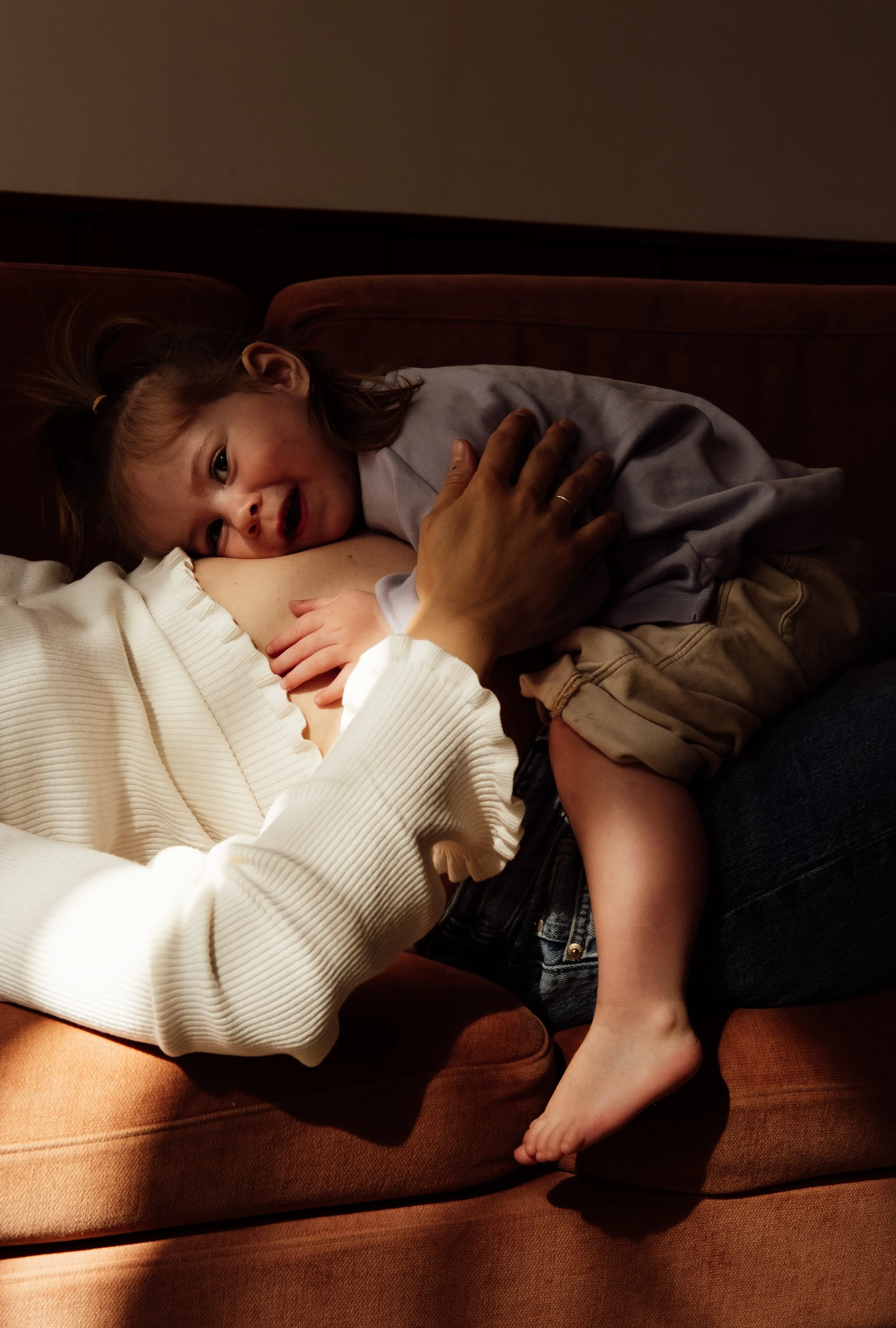 A young girl with a ponytail lying on a couch, smiling and cuddling with an adult. The adult's arm and hand are around the girl, and the girl has her head on the adult's arm. The scene appears warm and cozy in a dimly-lit room.