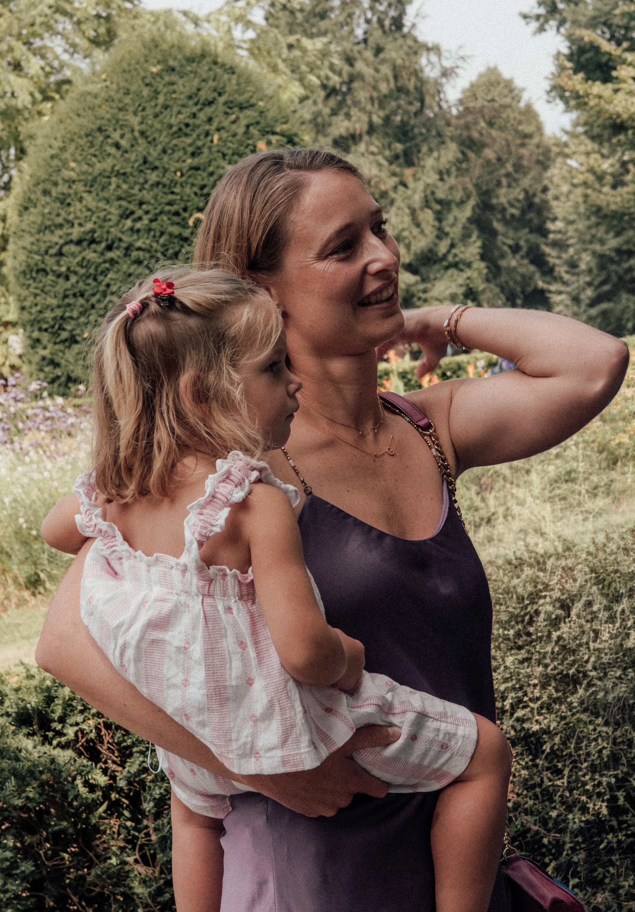 A woman holding a young girl outdoors, both smiling and enjoying a moment together with a background of green trees and bushes.