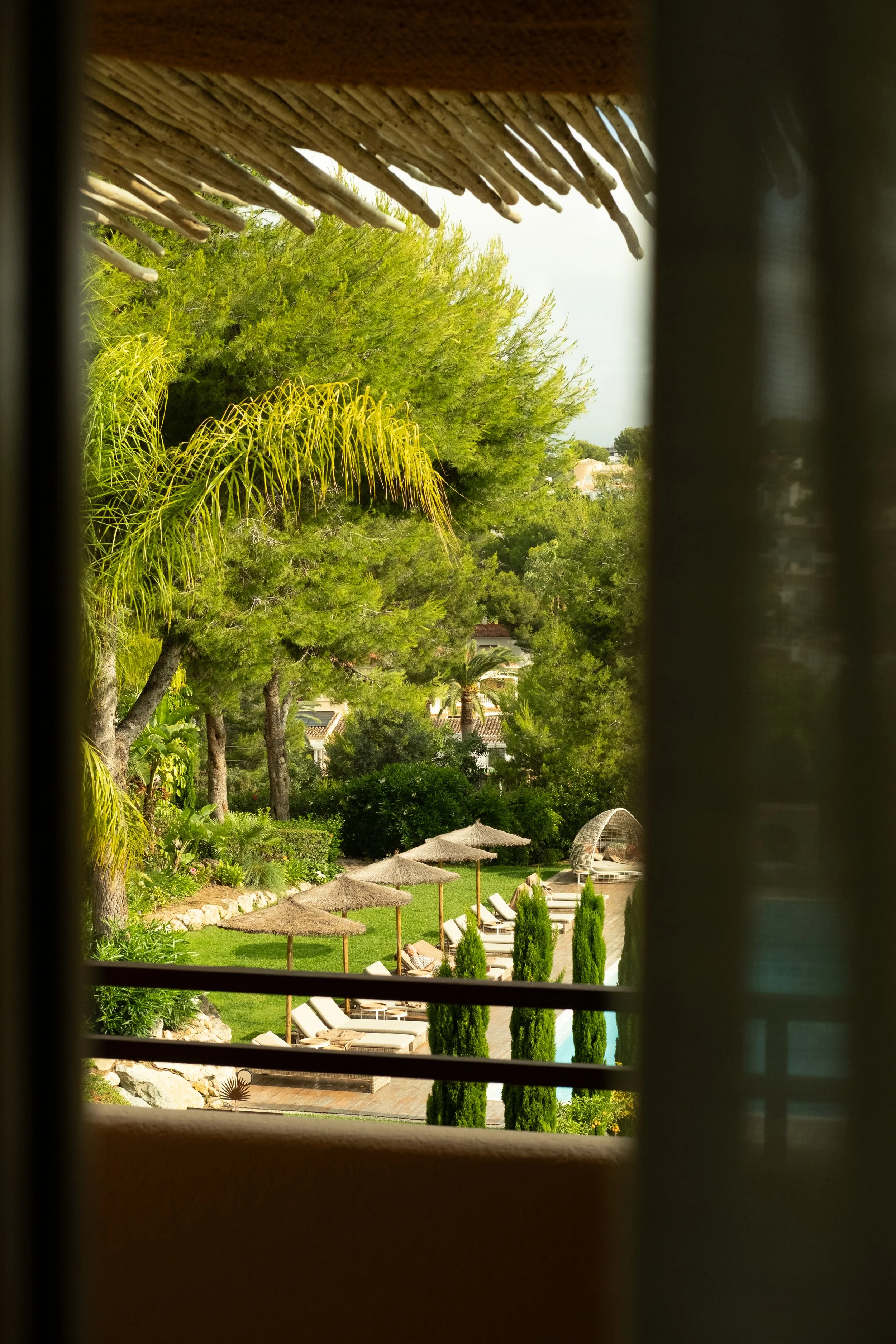 View of a lush green outdoor area through window shutters, with trees, a pool, and outdoor seating with straw umbrellas.