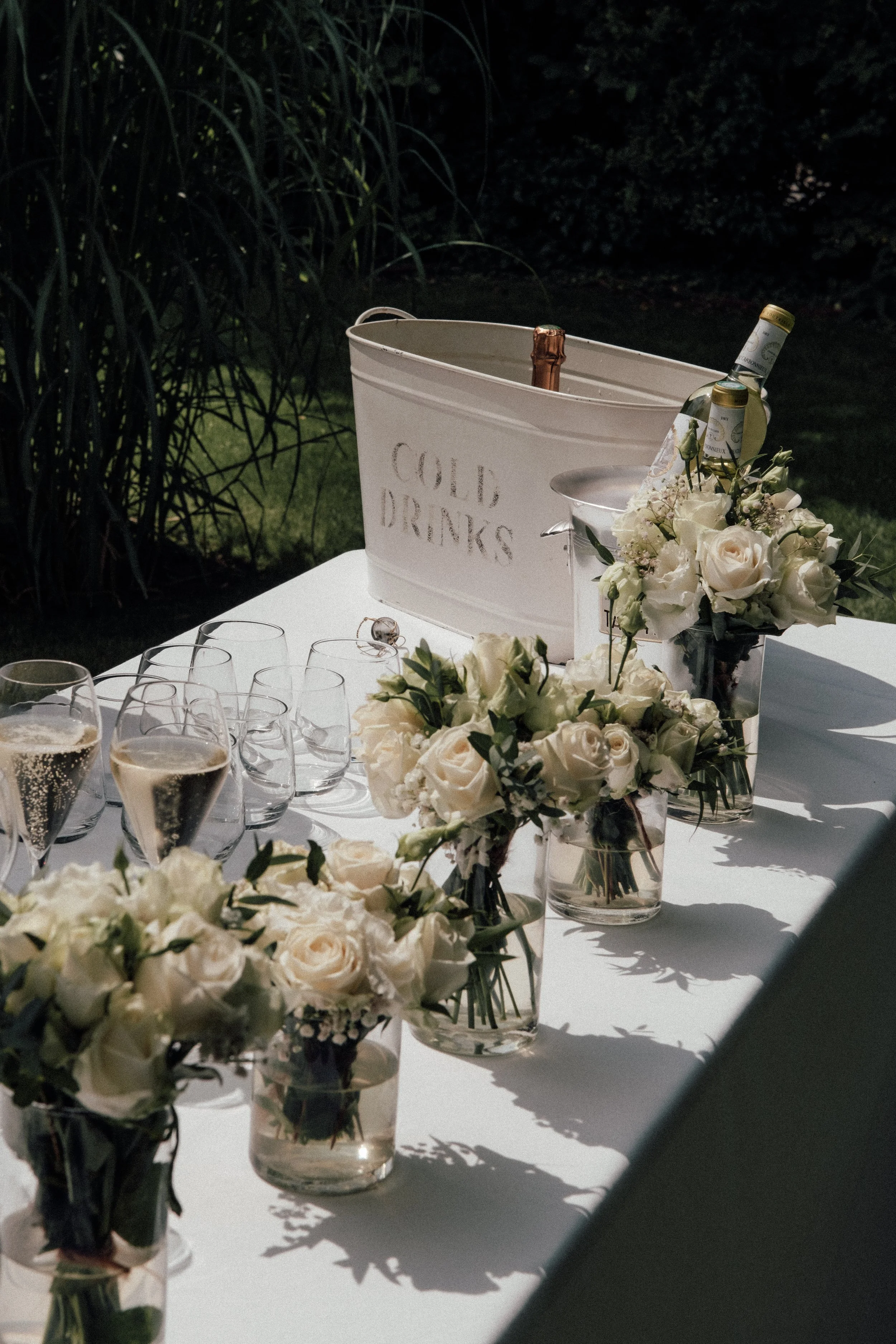 A table decorated with white roses in vases, glasses of champagne, and a white cooler with the words 'Cold Drinks' on it, set outdoors at night.