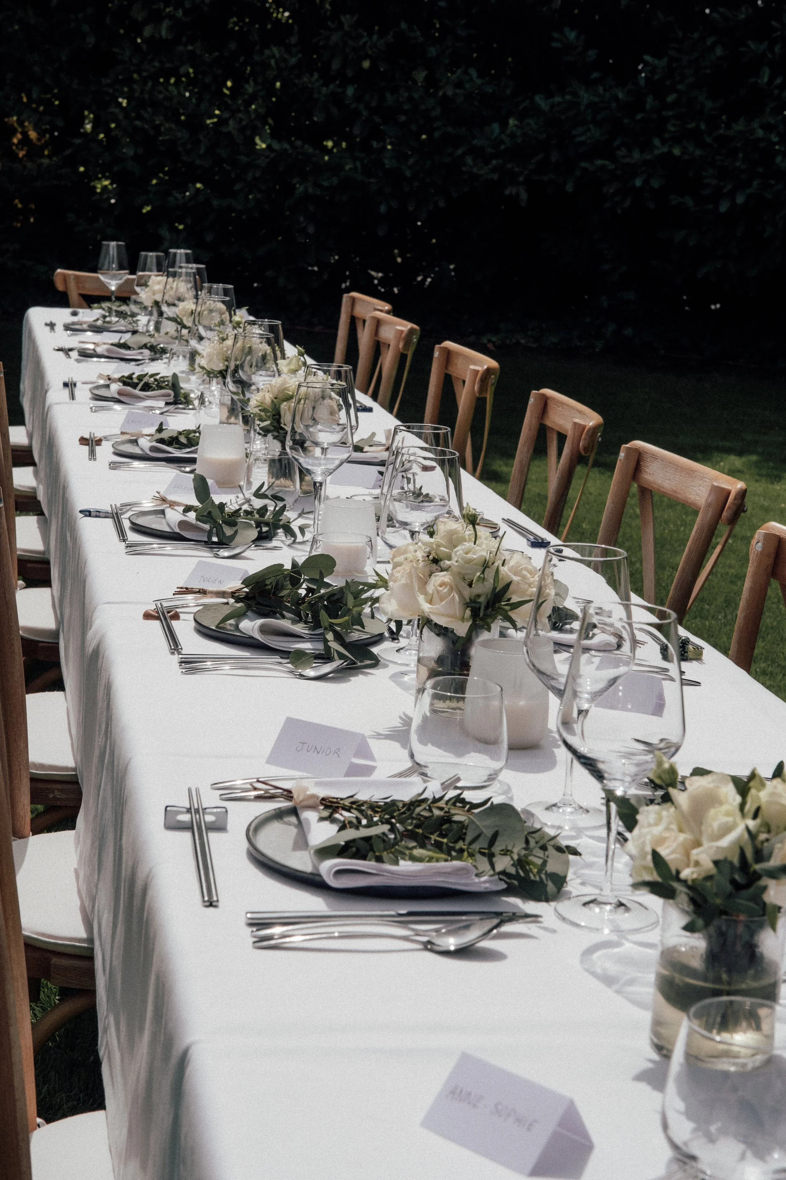 A long outdoor dining table set for a wedding or formal event, decorated with white flowers, greenery, and lit candles, surrounded by wooden chairs, with dark greenery in the background.