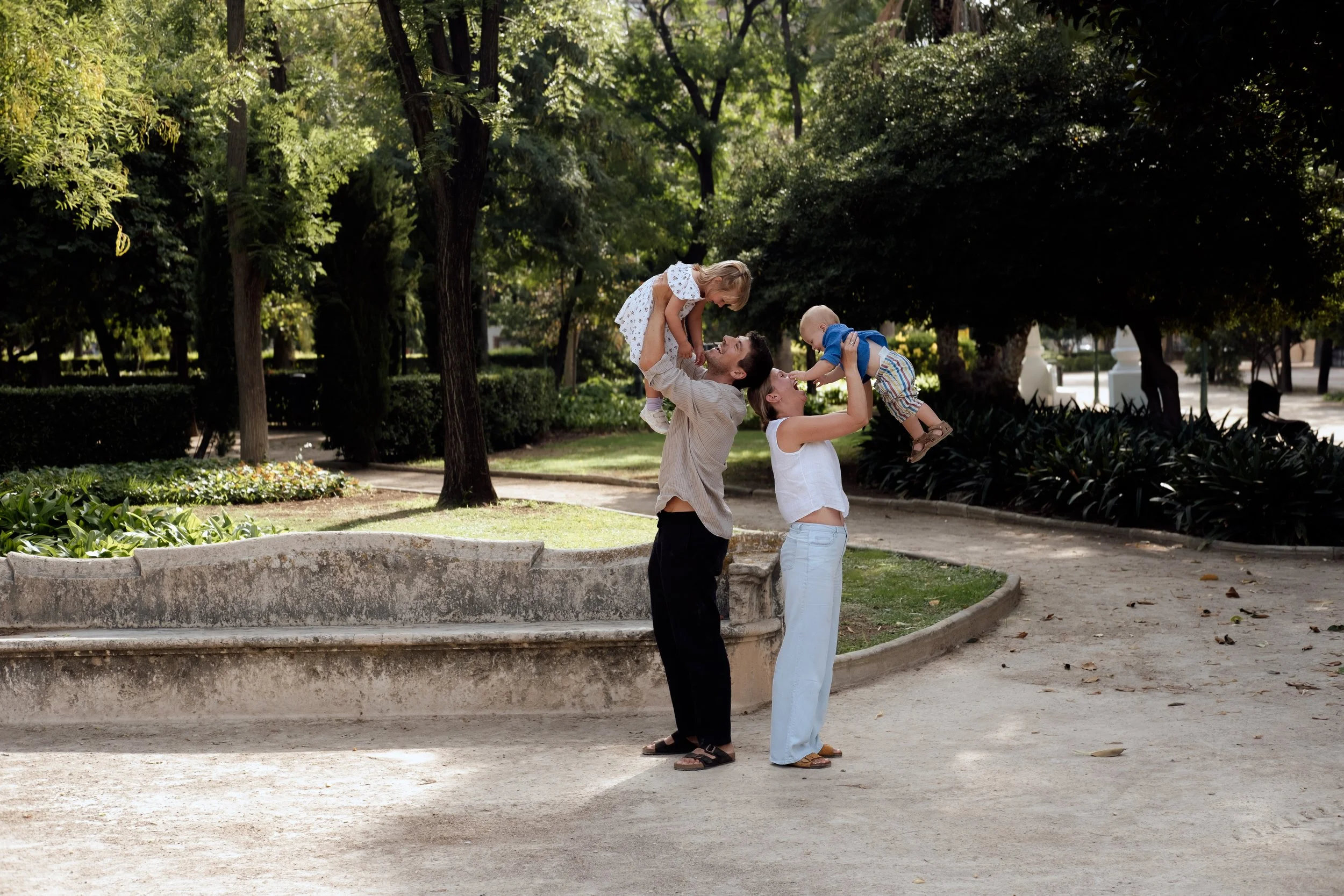 A family of four playing in a park, with two women lifting two children in the air, surrounded by trees and greenery.