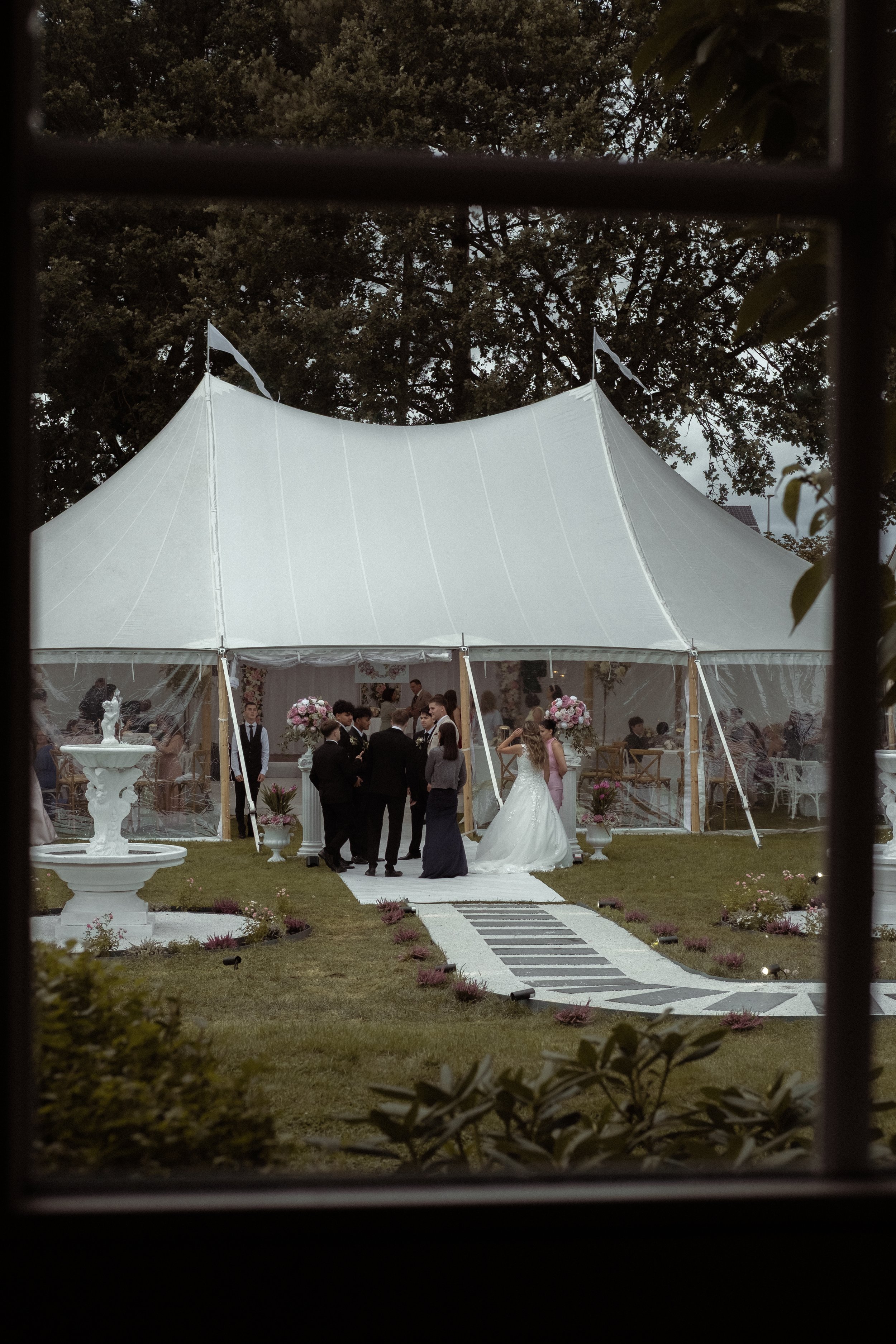 A wedding reception scene is visible through a window, showing a large white tent, guests socializing, and a bride in a wedding dress walking with others outside.