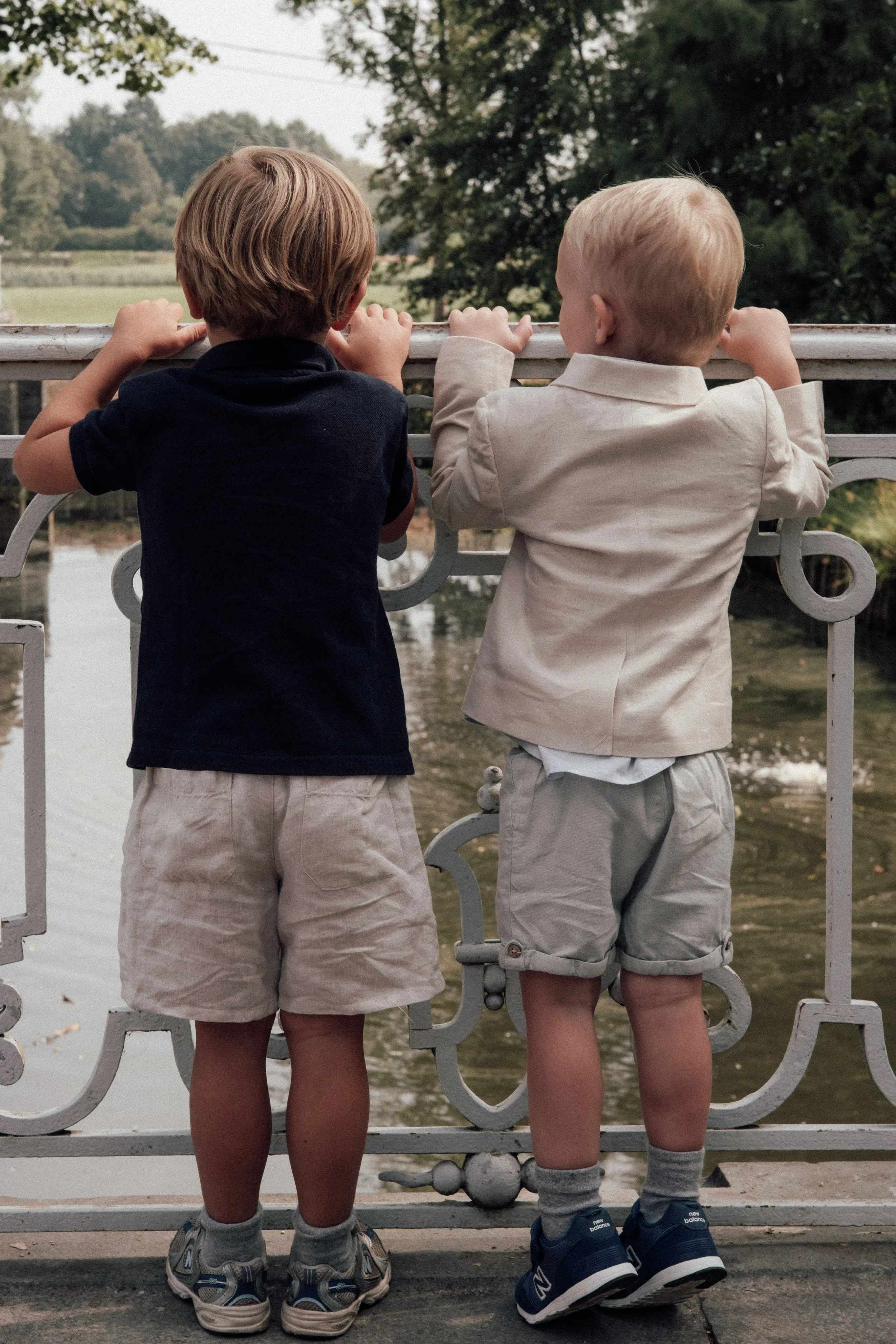 Two young boys standing side by side on a bridge, looking out over a river and lush greenery, with their backs to the camera.