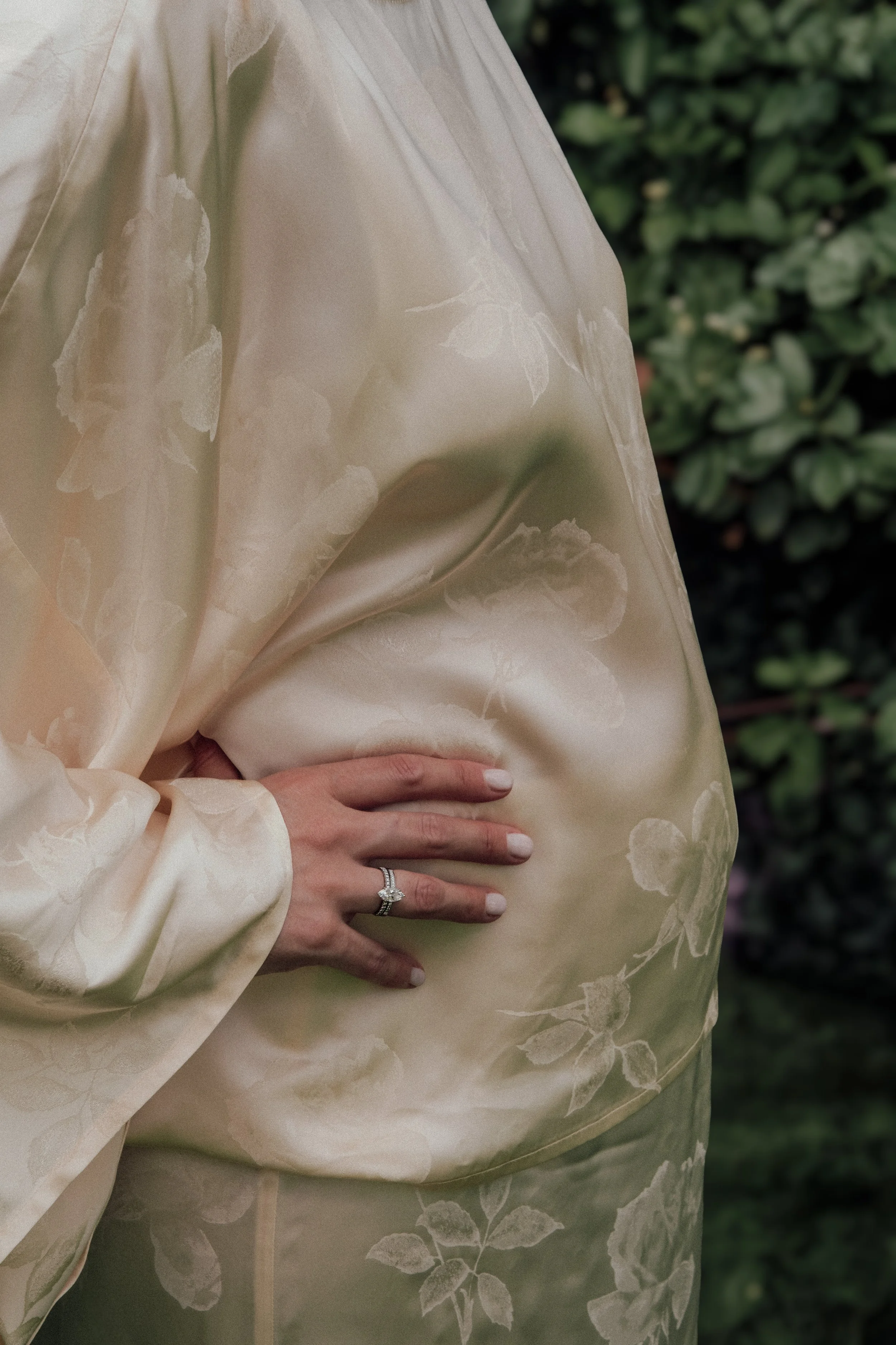 Person wearing a cream-colored dress with floral patterns, holding their waist with their left hand, which has a silver ring with a large gemstone.