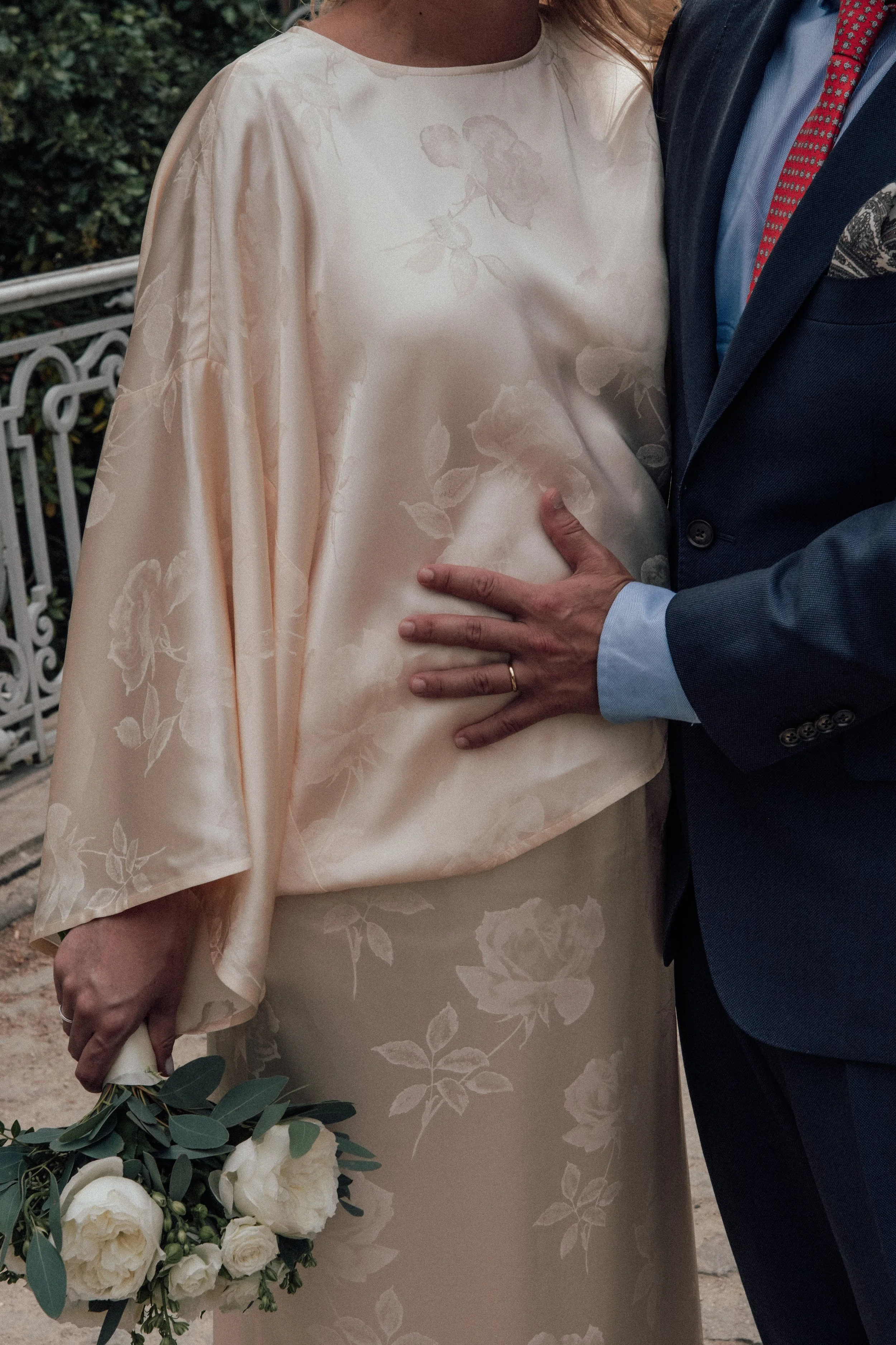 A close-up of a bride and groom during a wedding, with the bride wearing a cream-colored floral dress and holding a bouquet of white roses and greenery, and the groom wearing a dark blue suit with a red patterned tie. The groom's hand is on the bride