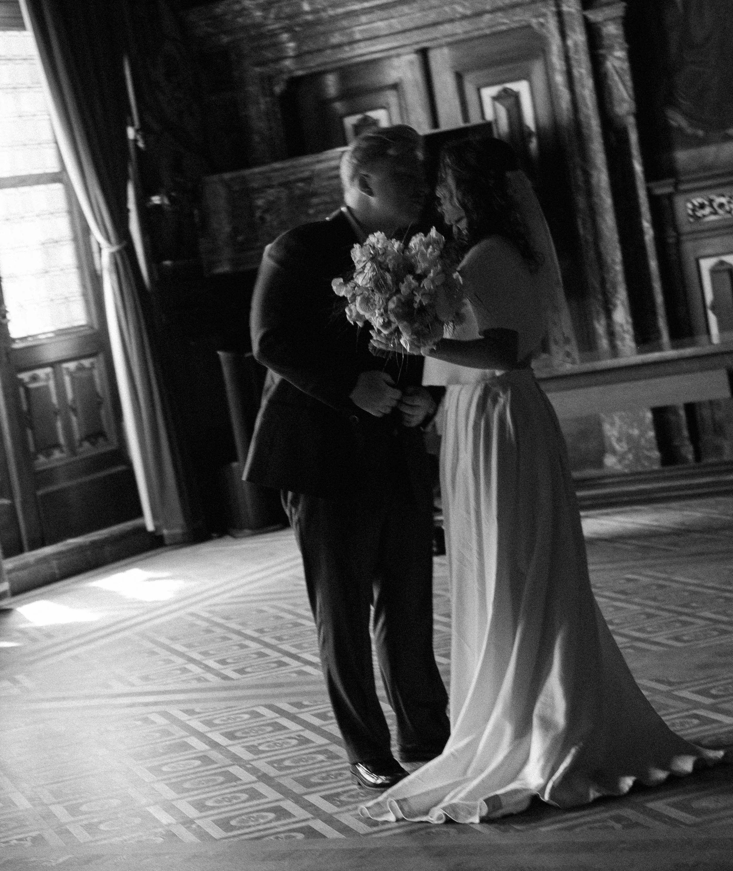 A couple shares a kiss in a wedding dress and suit, standing close together in front of a mirror, in a vintage-style room with wooden furniture and patterned flooring.