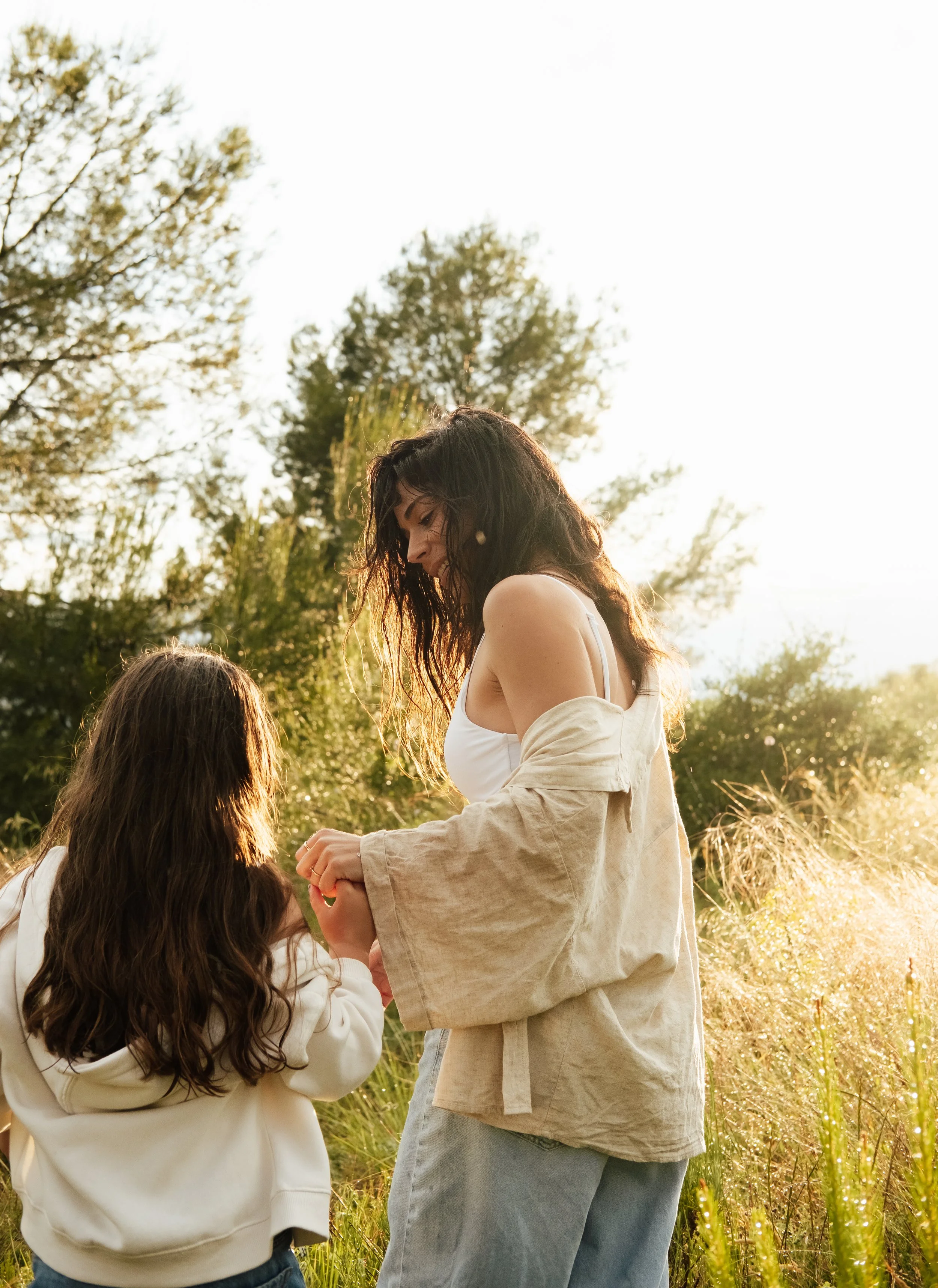 A woman with curly dark hair in a beige oversized jacket and a white dress, smiling down at a young girl with long brown hair, who is holding her hands together, in a grassy field with trees in the background, during golden hour sunlight.