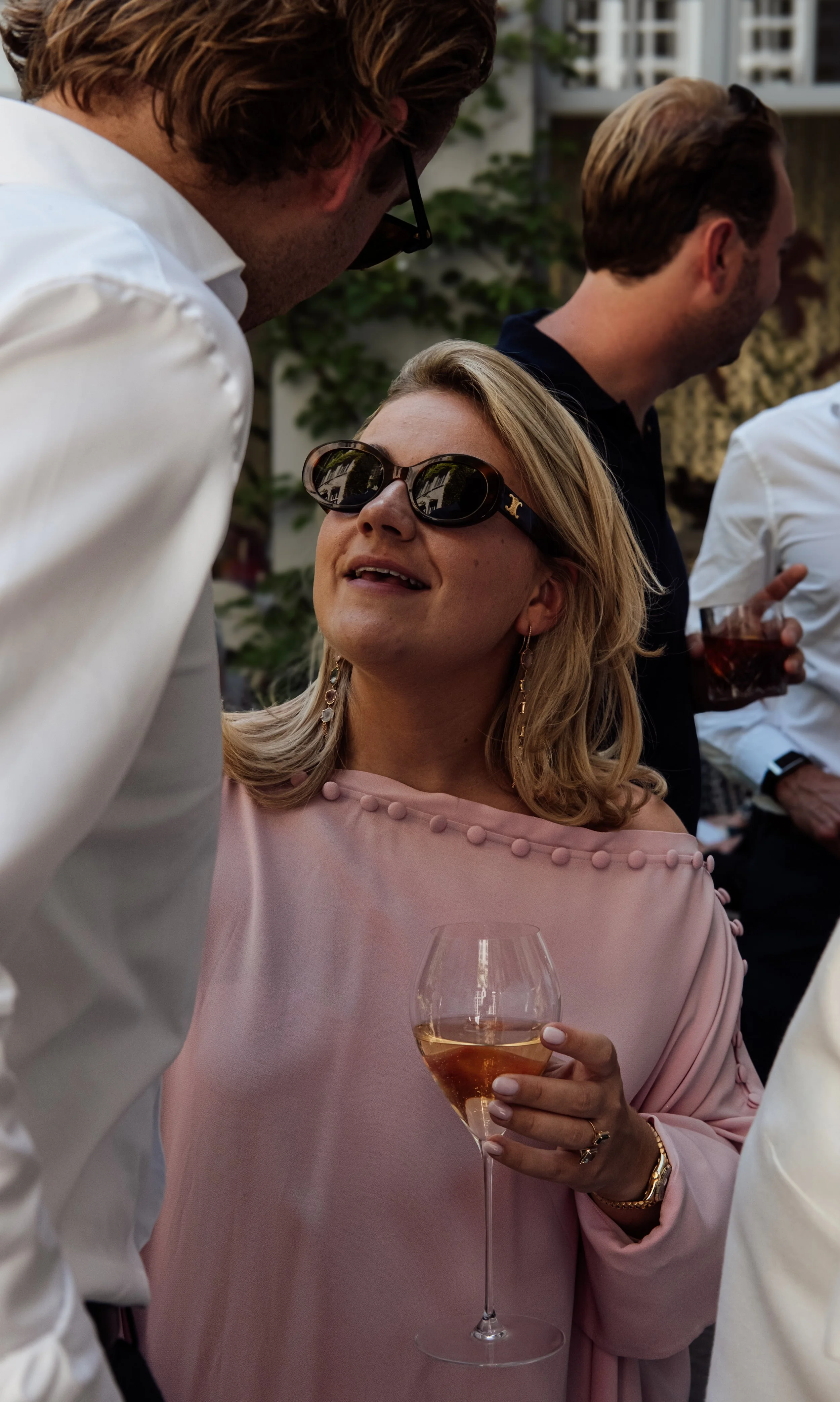 A woman in pink dress holding a glass of rosé wine at a social gathering, talking to a man with glasses leaning in close, with other people in the background.