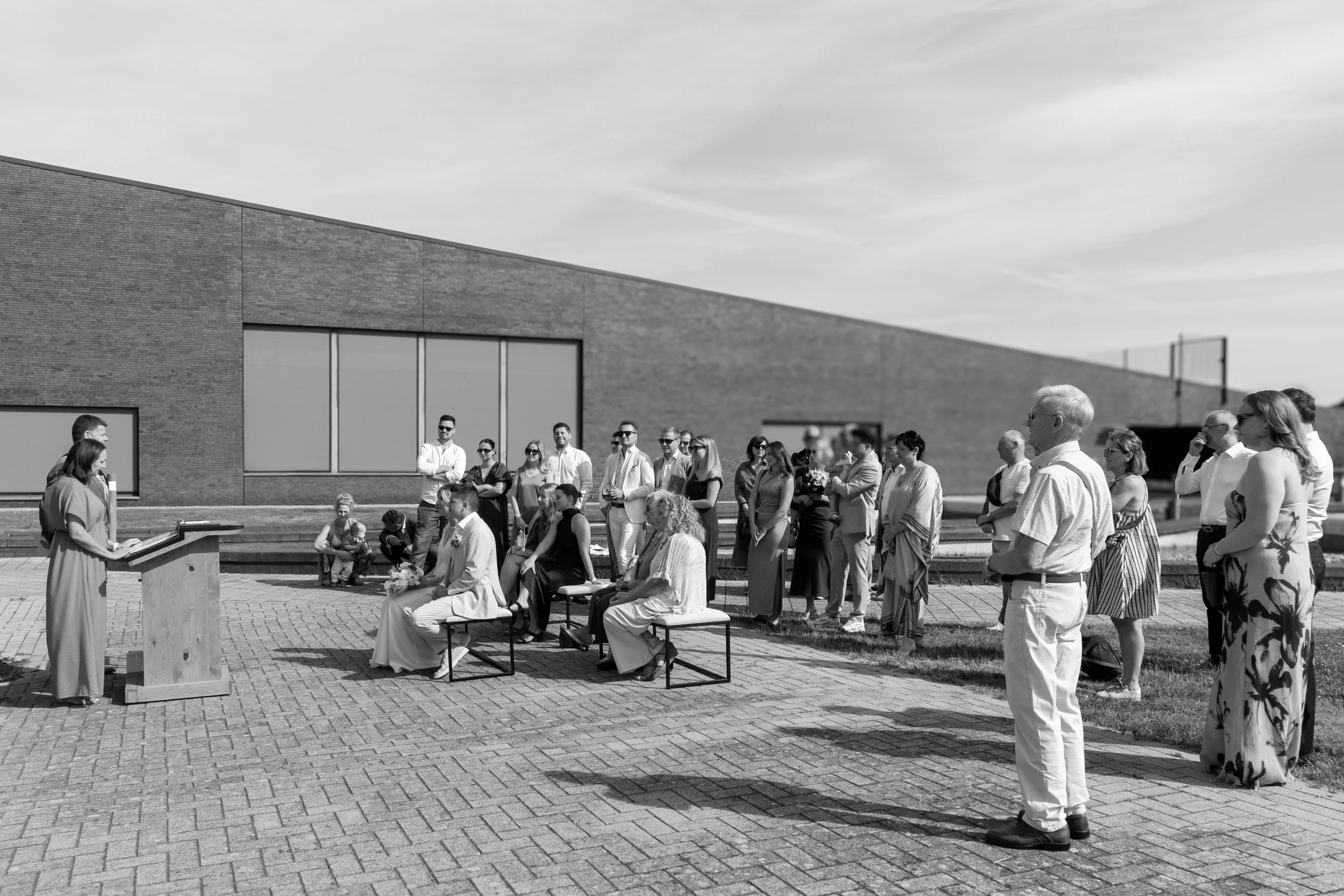 A black and white photo of a group of people gathered outdoors near a modern brick building, attending an event with a woman reading at a podium.