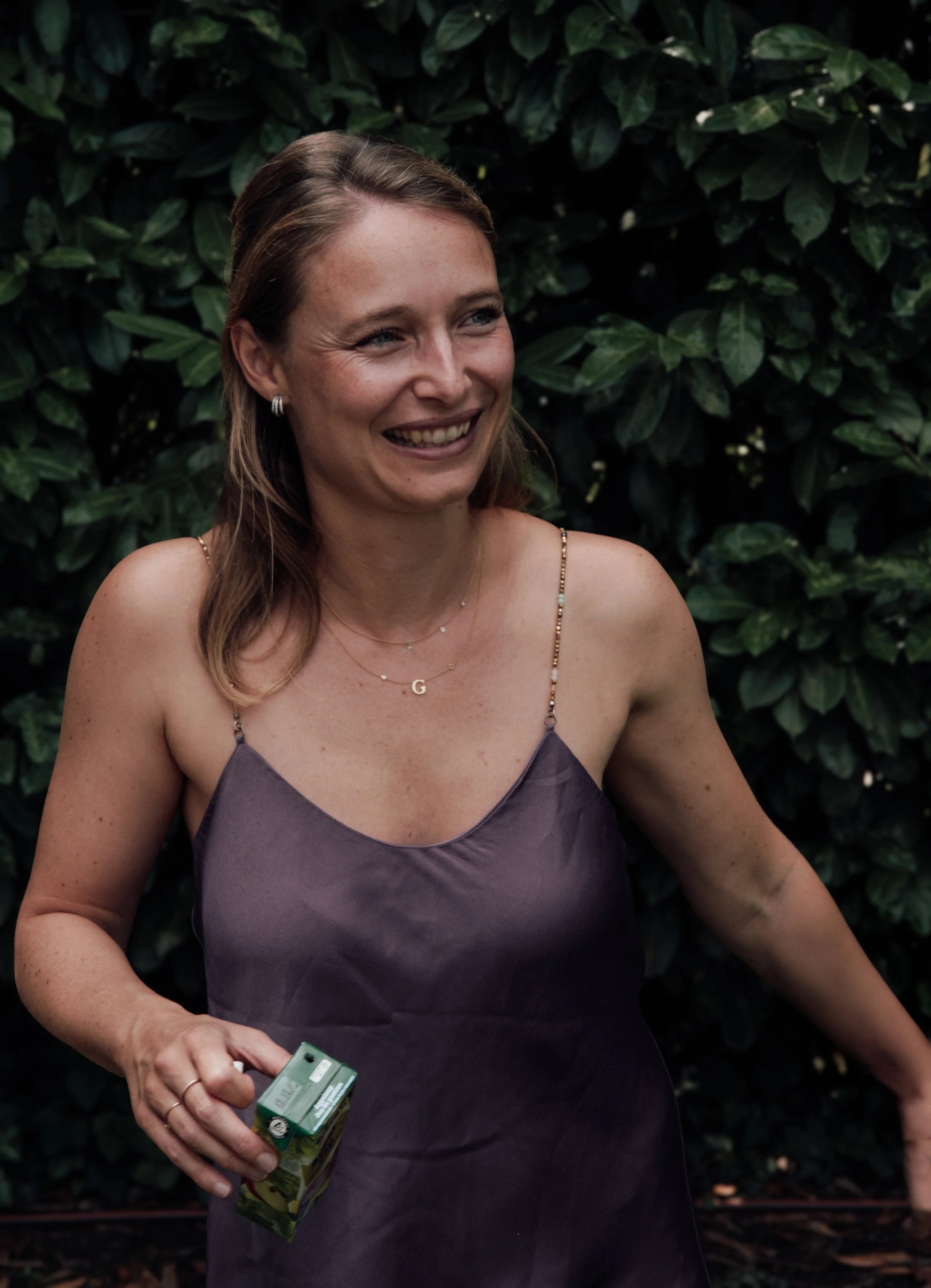 A woman smiling and holding a small carton in her right hand, standing in front of green leafy bushes.