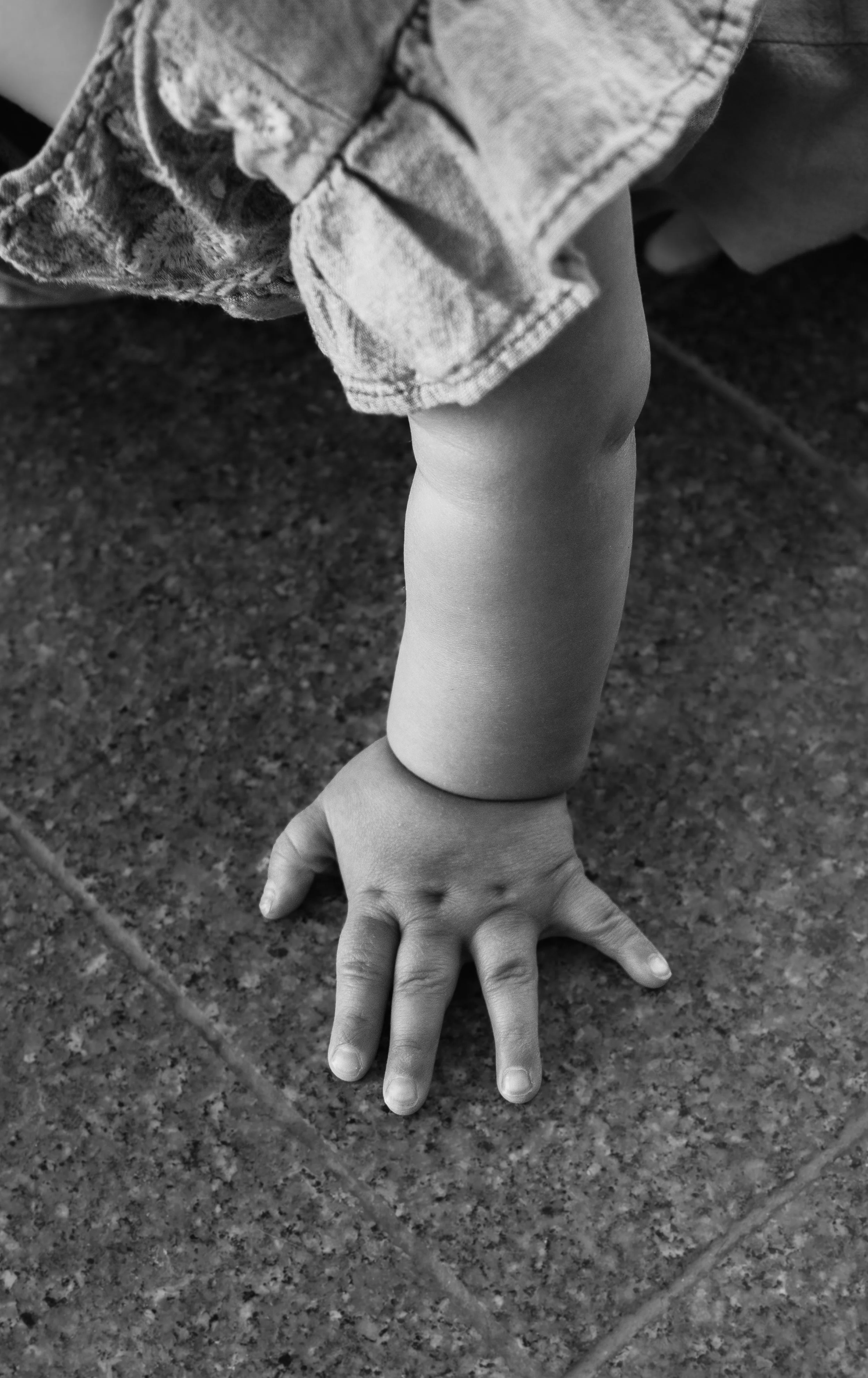 A child's hand and arm stretched out on a tiled floor, with the child's body and a sleeve visible in the top portion of the image.