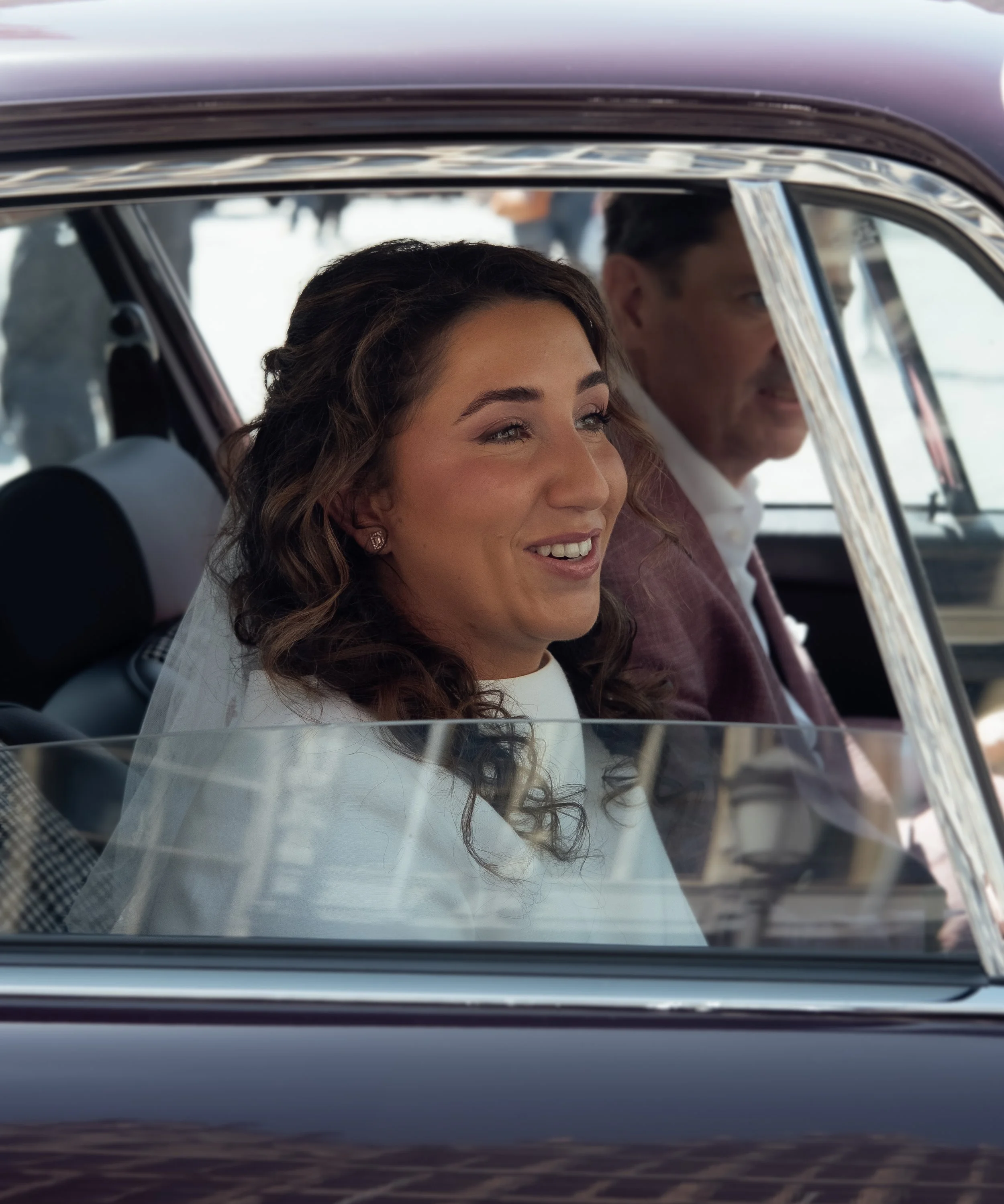A woman with curly brown hair and earrings sitting in the passenger seat of a car, smiling, with a man with dark hair and a beard sitting in the driver seat, both viewed through the car's window.