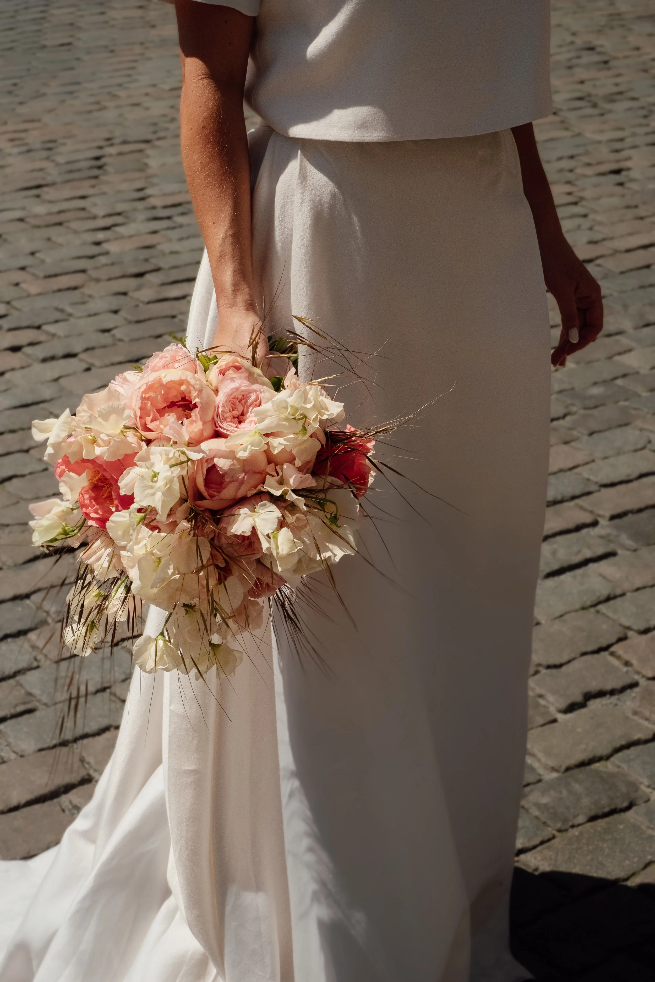 Close-up of a person in a white dress holding a pink and white floral bouquet with cobblestone pavement in the background.