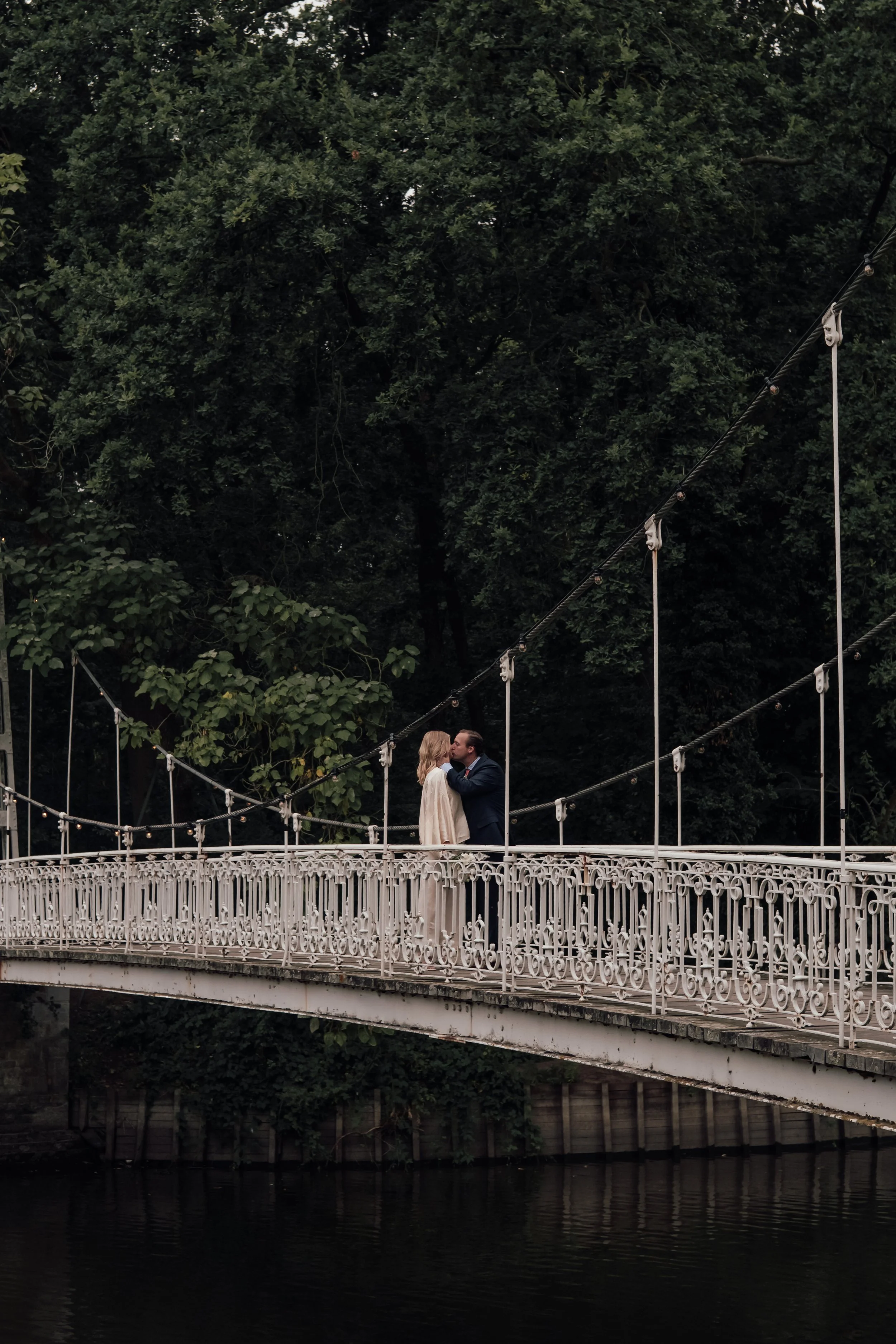 A couple sharing a kiss on a Victorian-style pedestrian bridge over water, surrounded by dense green trees.