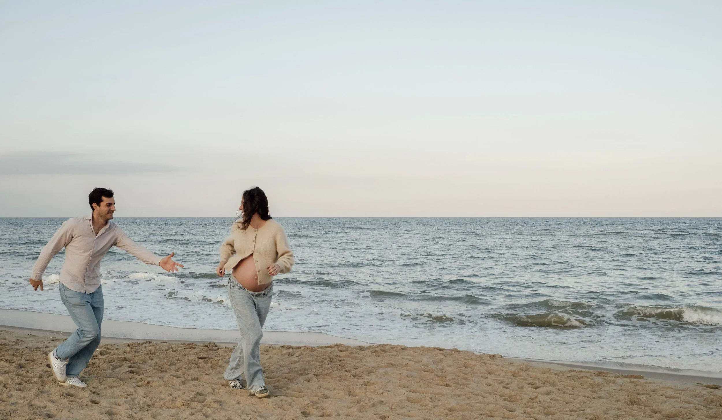 A man and a pregnant woman enjoying time at the beach, holding hands and smiling, with ocean waves and a cloudy sky in the background.