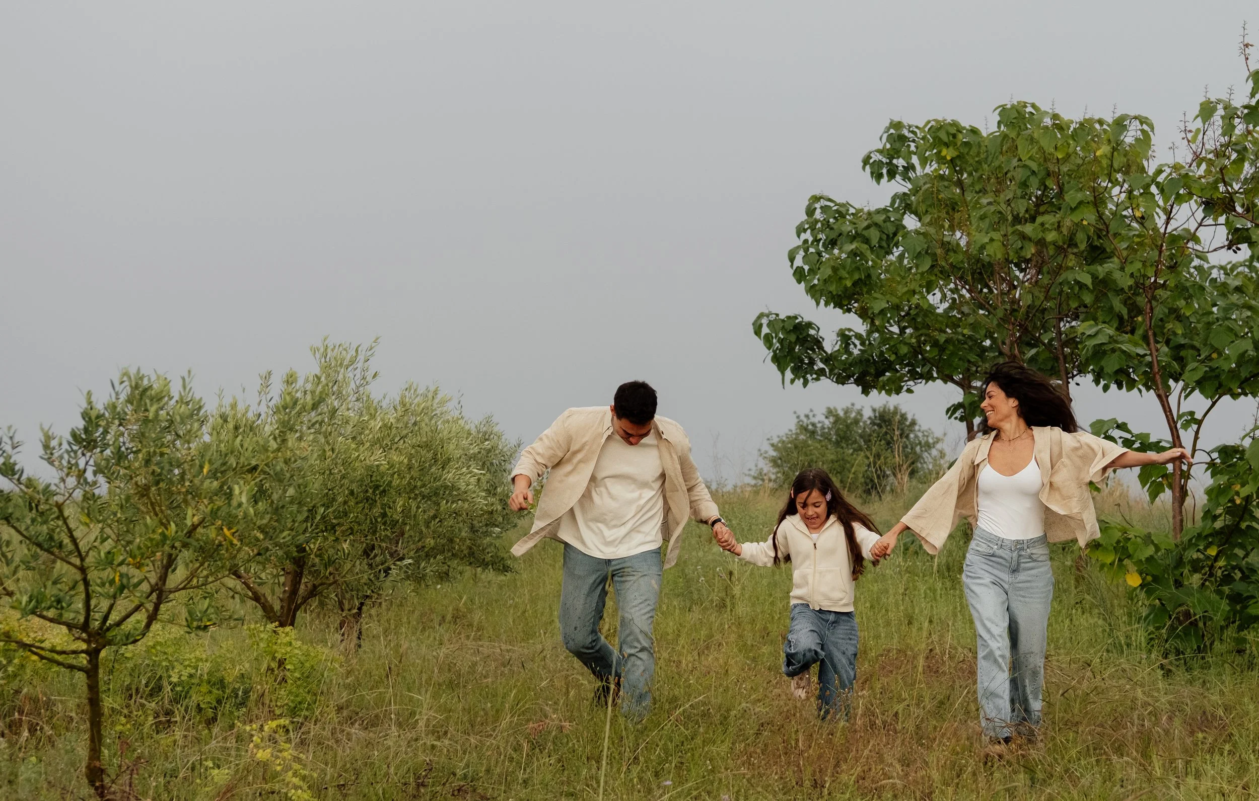 A family of four, including a man, woman, and two young girls, holding hands and walking joyfully through a green field with trees, under a cloudy sky.