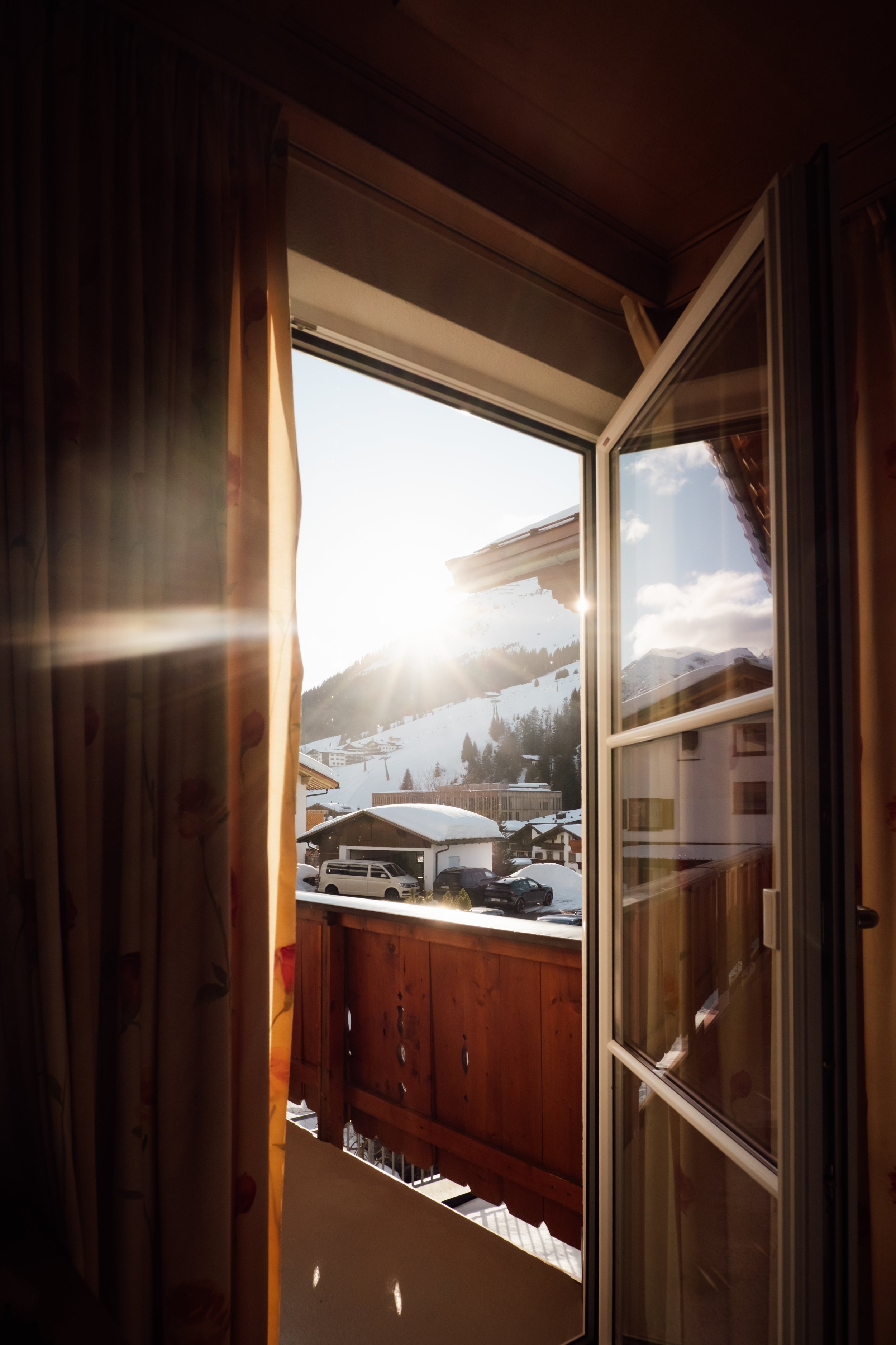 Sunset view from an open door onto a snowy mountain landscape with parked cars and buildings in the foreground.