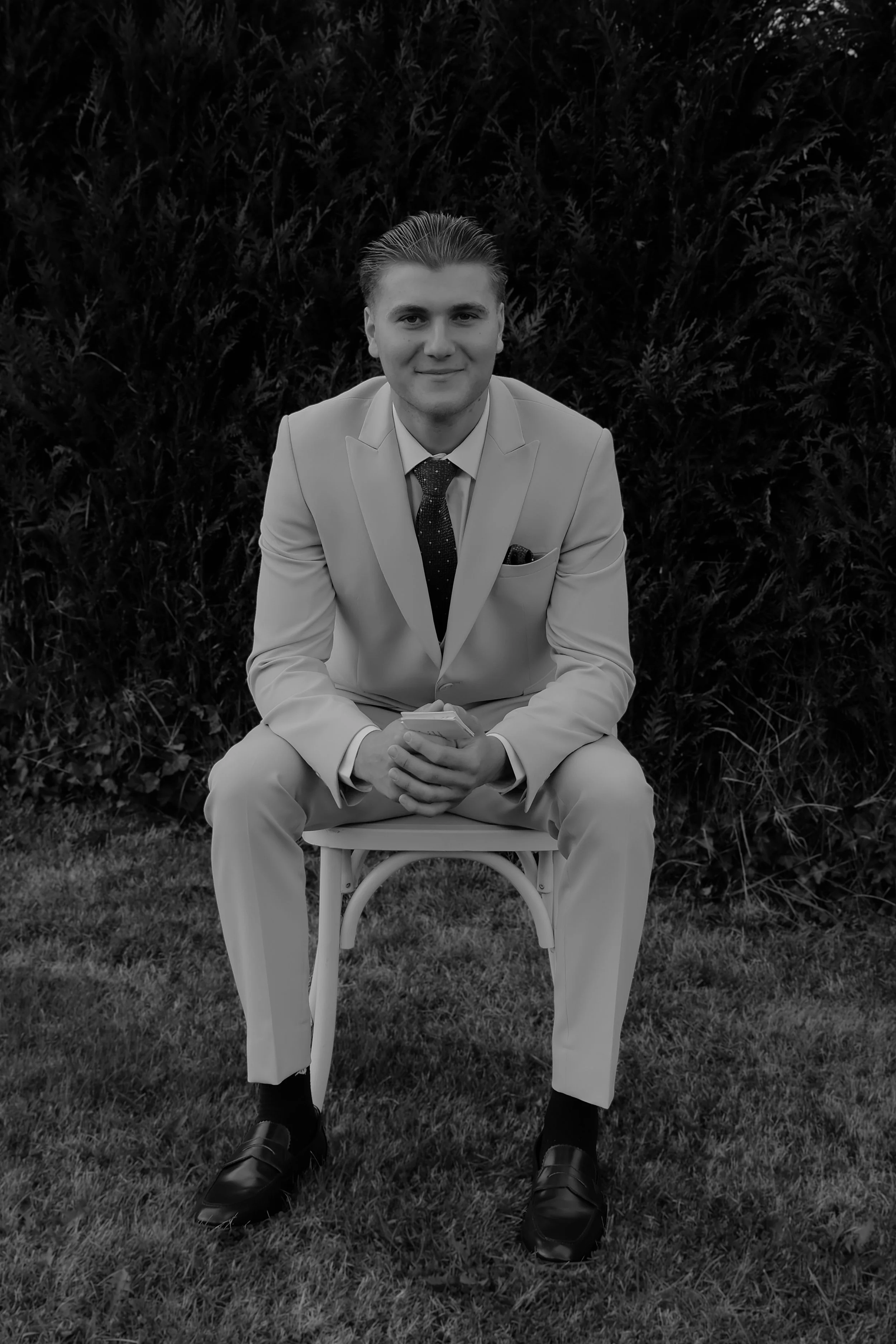 A black and white photo of a young man with slicked-back hair, dressed in a suit with a tie and pocket square, sitting on a white chair outdoors against a background of bushes. He is smiling slightly and holding a phone.