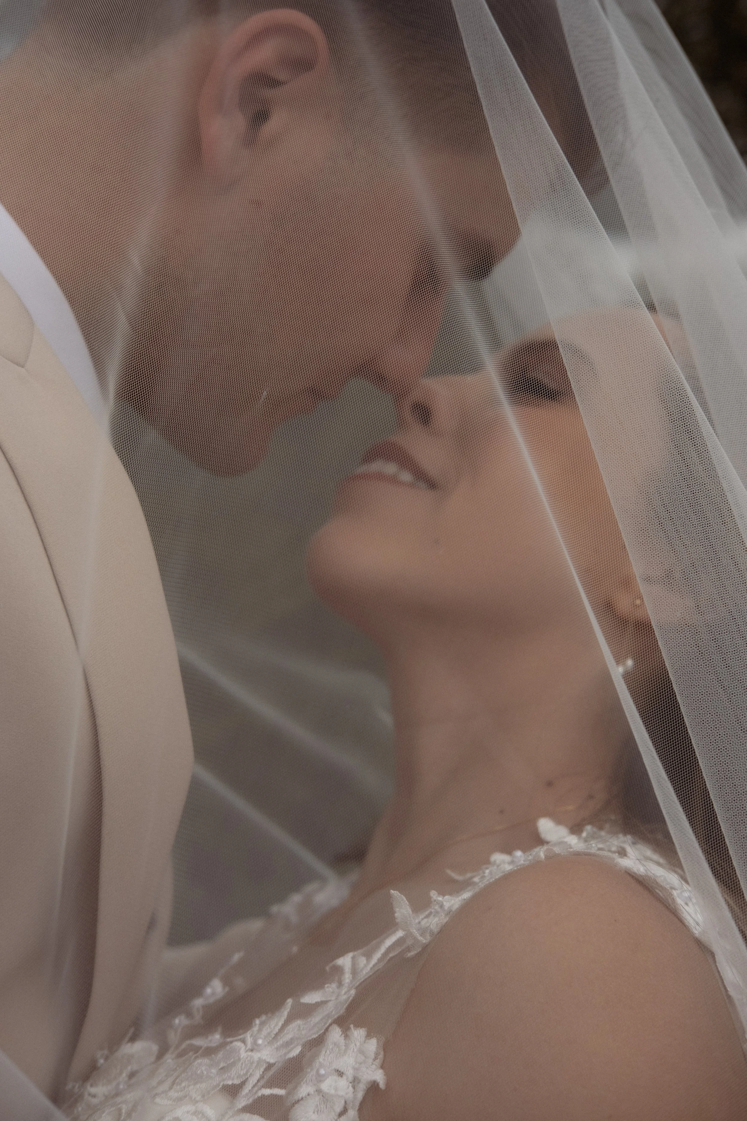 A close-up of a bride with her eyes closed, smiling softly under a veil, wearing a white dress with lace details, captured in a romantic moment.