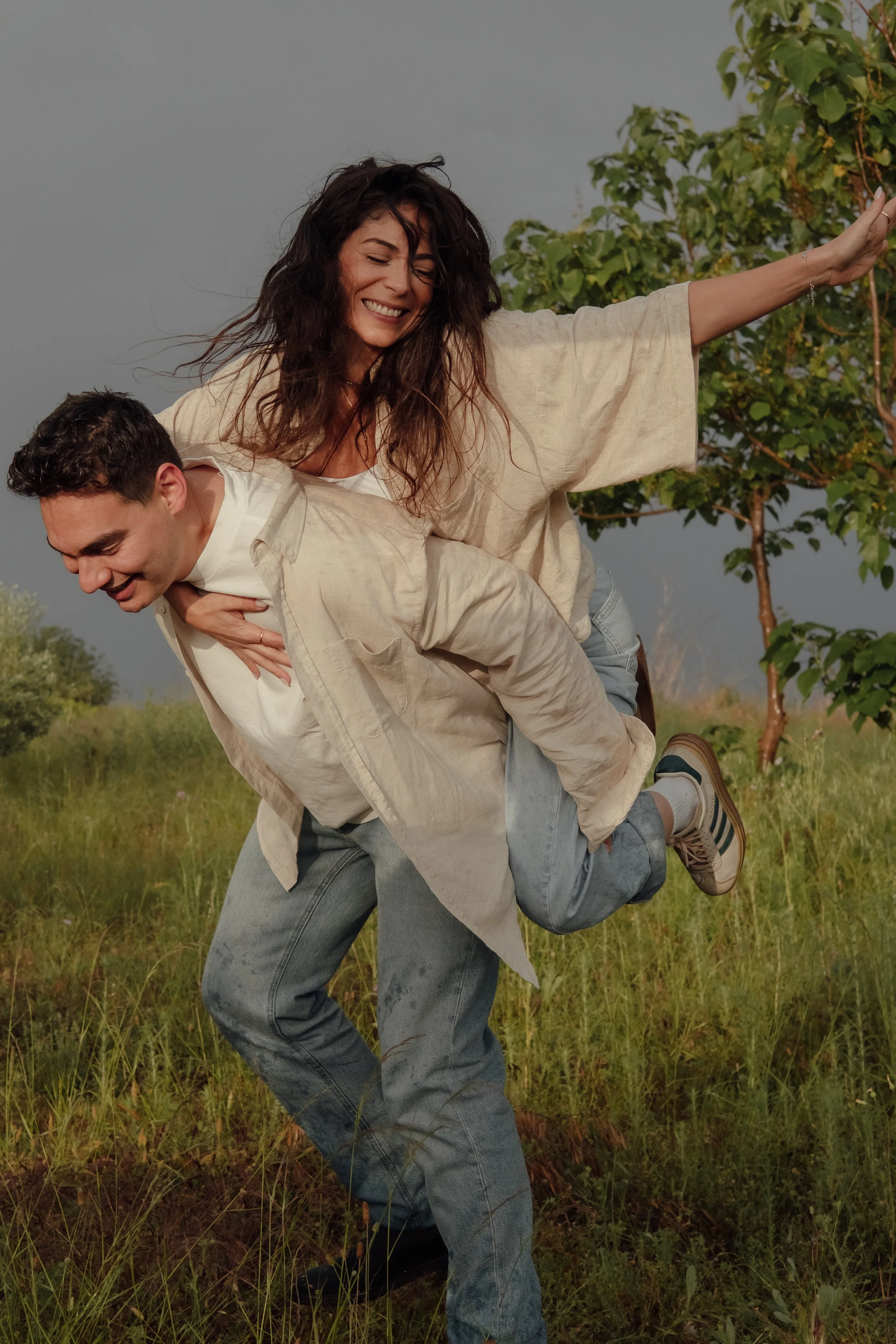 A woman smiling and laughing while sitting on a man's shoulders in a grassy outdoor setting, with trees in the background.