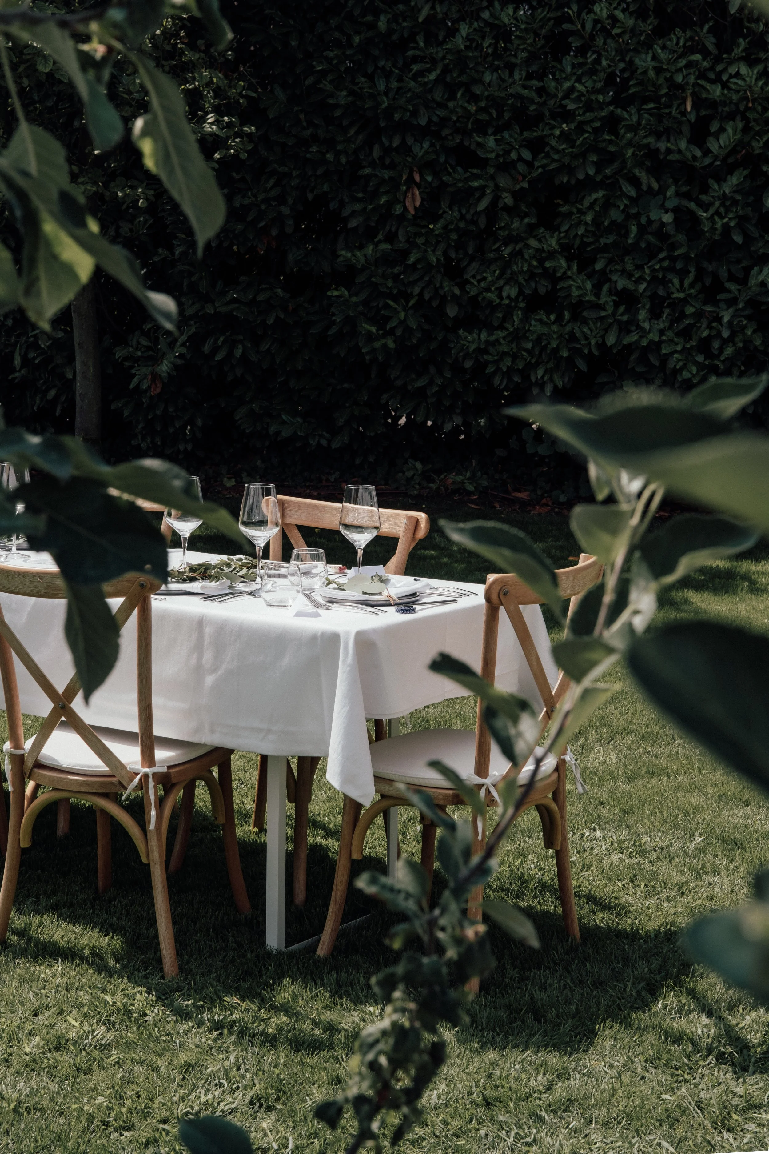 Garden outdoor dining setup with a round table covered with a white tablecloth, set with wine glasses, silverware, and napkins, surrounded by wooden chairs, and lush green hedge in the background.
