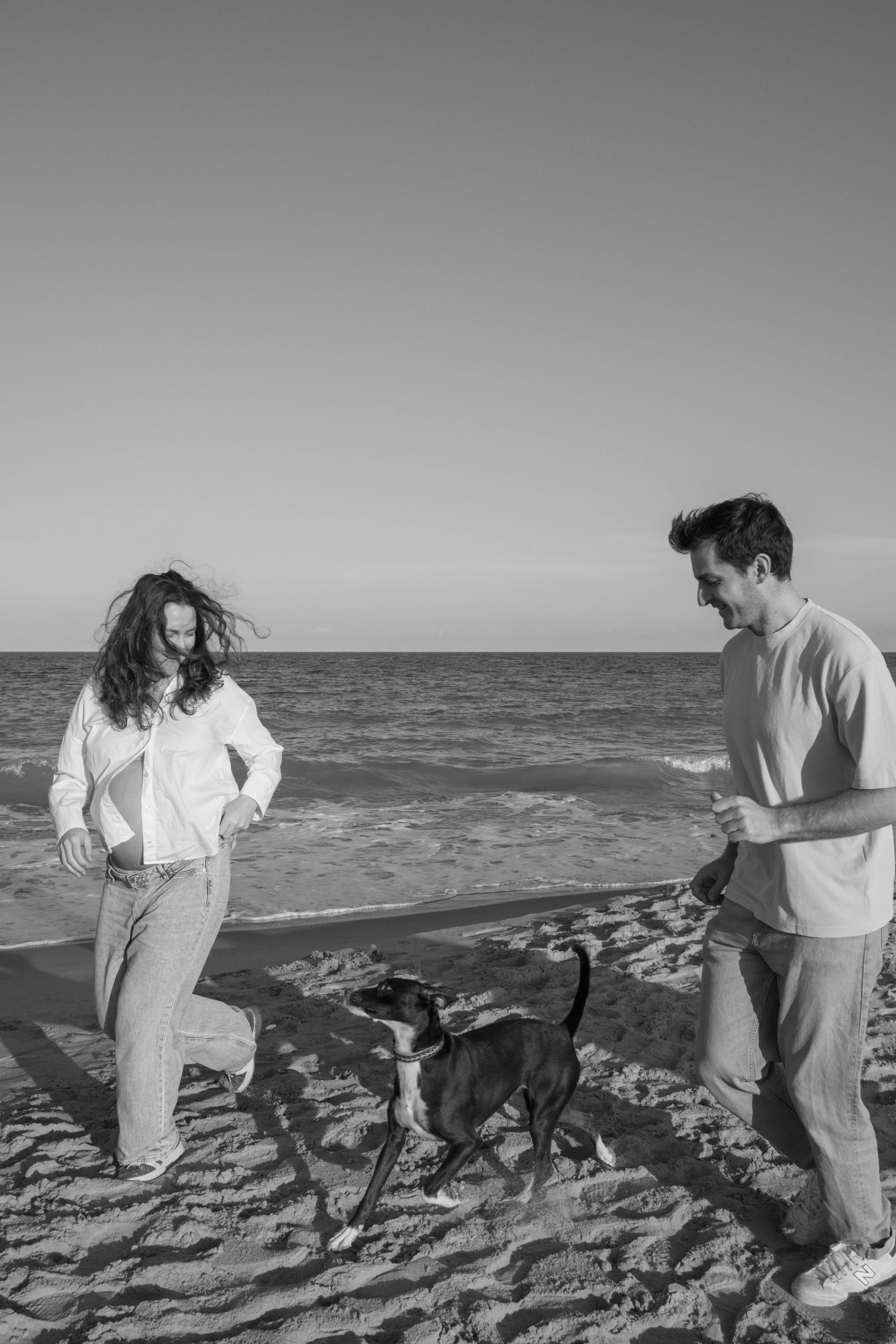 Two people and a dog on a beach, with the ocean and sky in the background. The woman on the left is wearing a white shirt and light jeans, and the man on the right is wearing a light-colored shirt and pants. The woman is looking down, while the man i