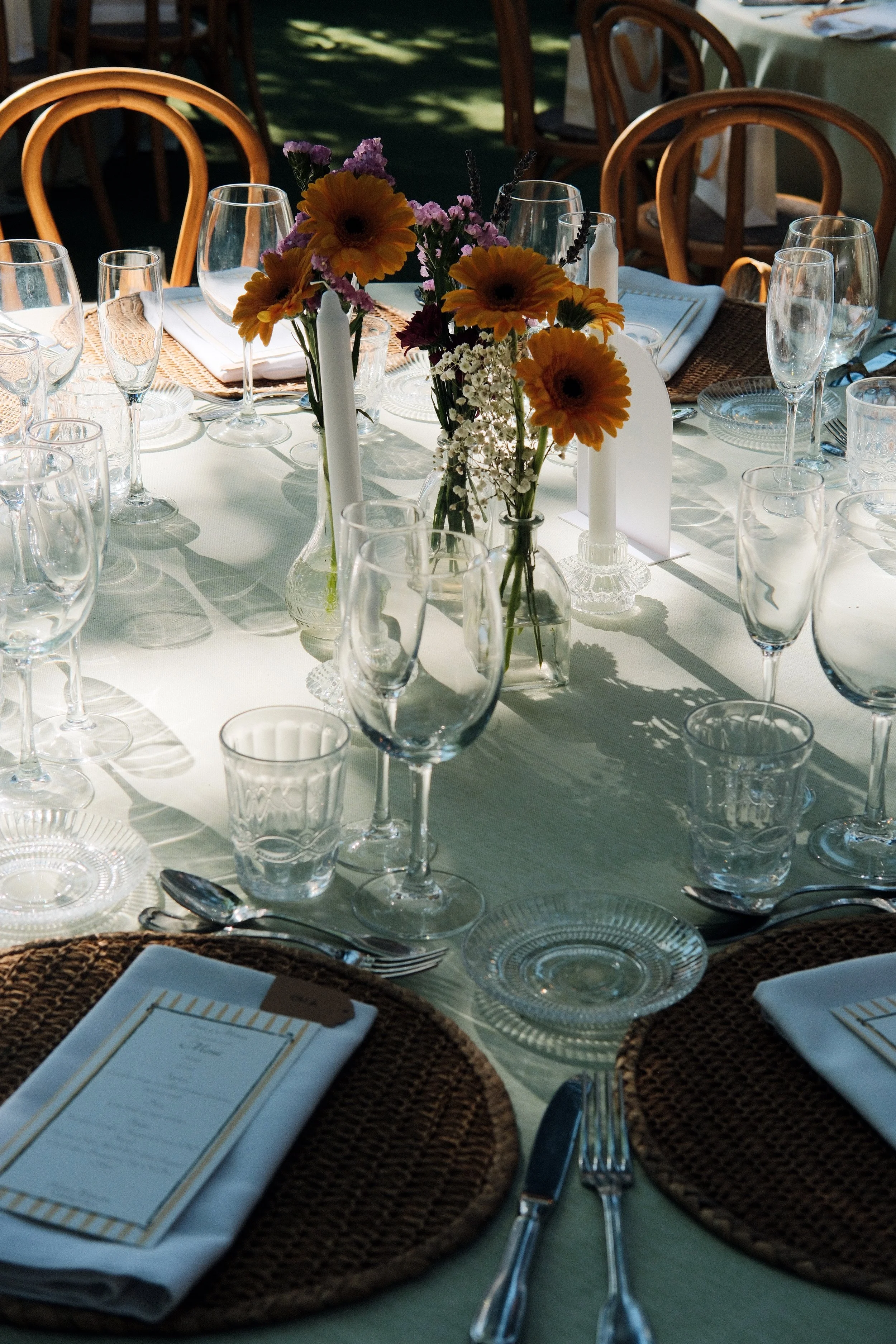 Elegant outdoor dining table set with glassware, floral centerpiece, and place settings, illuminated by sunlight.