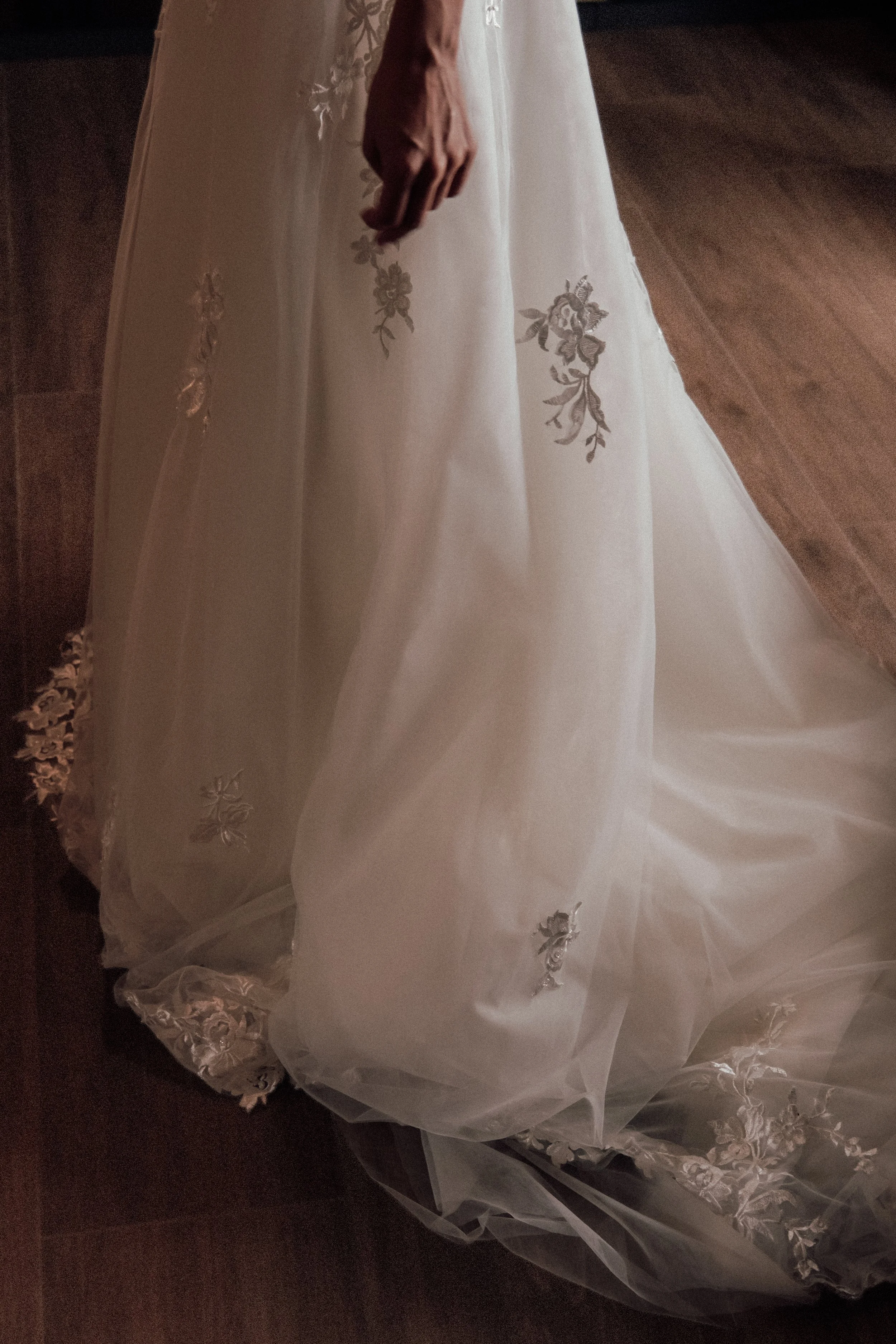 Close-up of a white wedding dress with floral embroidery, laying on a dark wooden floor.