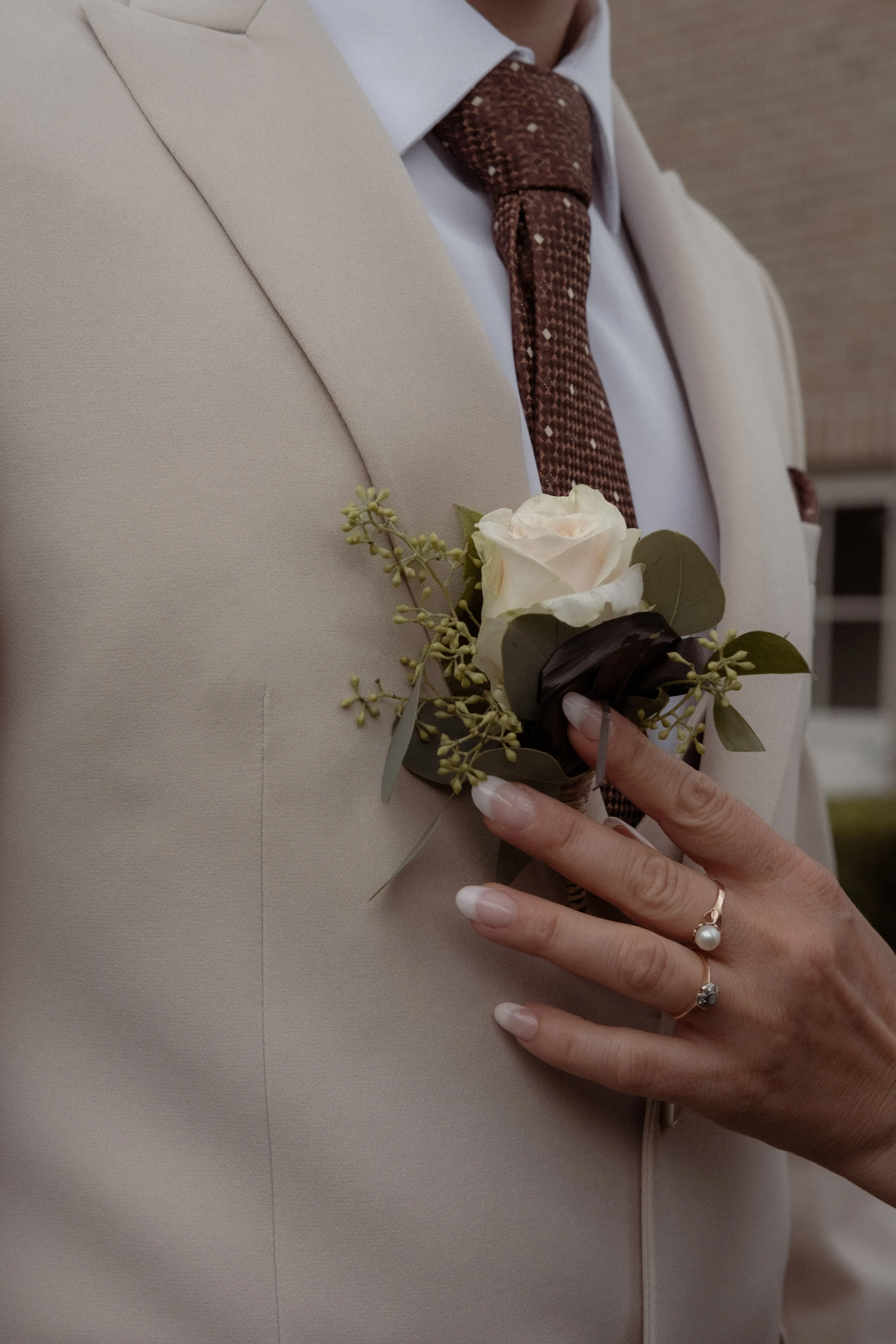 Close-up of a person in a white suit jacket holding a boutonniere with a white rose, eucalyptus, and other greenery, wearing rings.