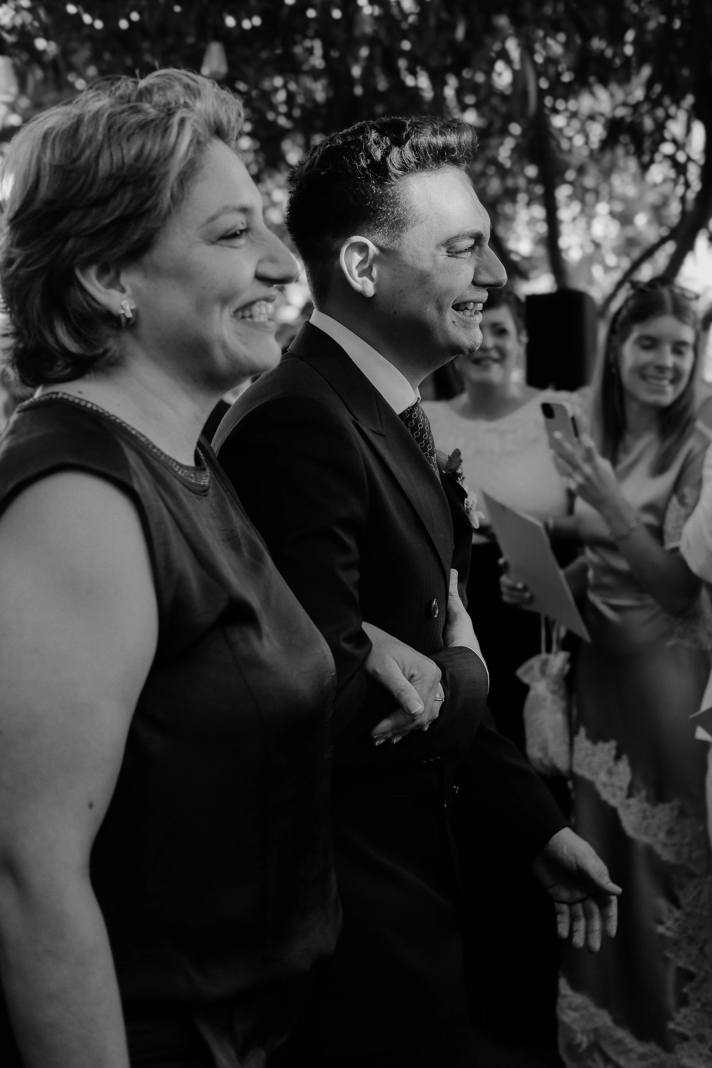 Black and white photo of multiple people smiling and sitting in a row outdoors, with trees in the background.