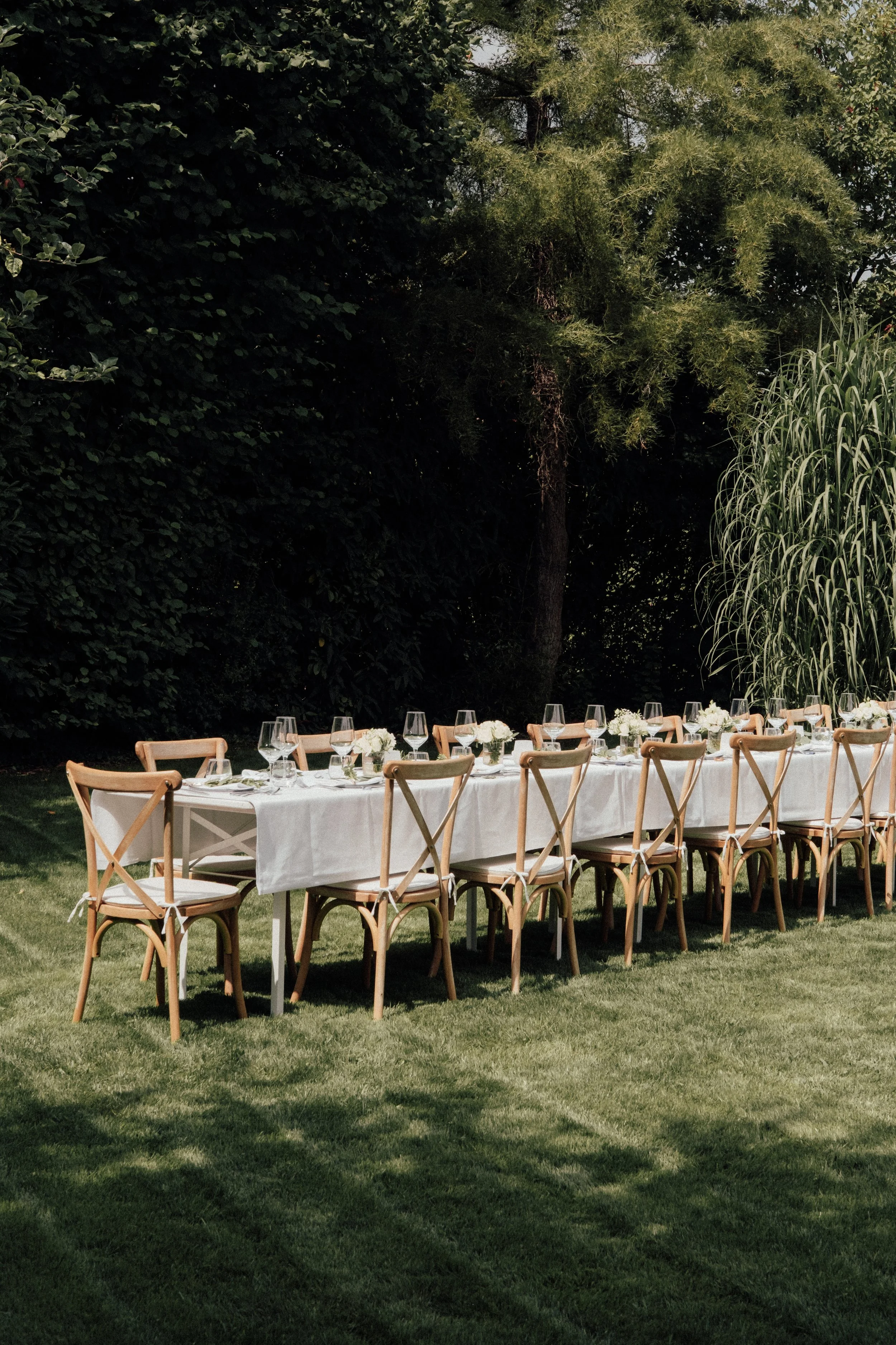 An outdoor long dining table set for a meal, with white tablecloths, floral centerpieces, wine glasses, and chairs, situated on a grassy area surrounded by trees and shrubs.