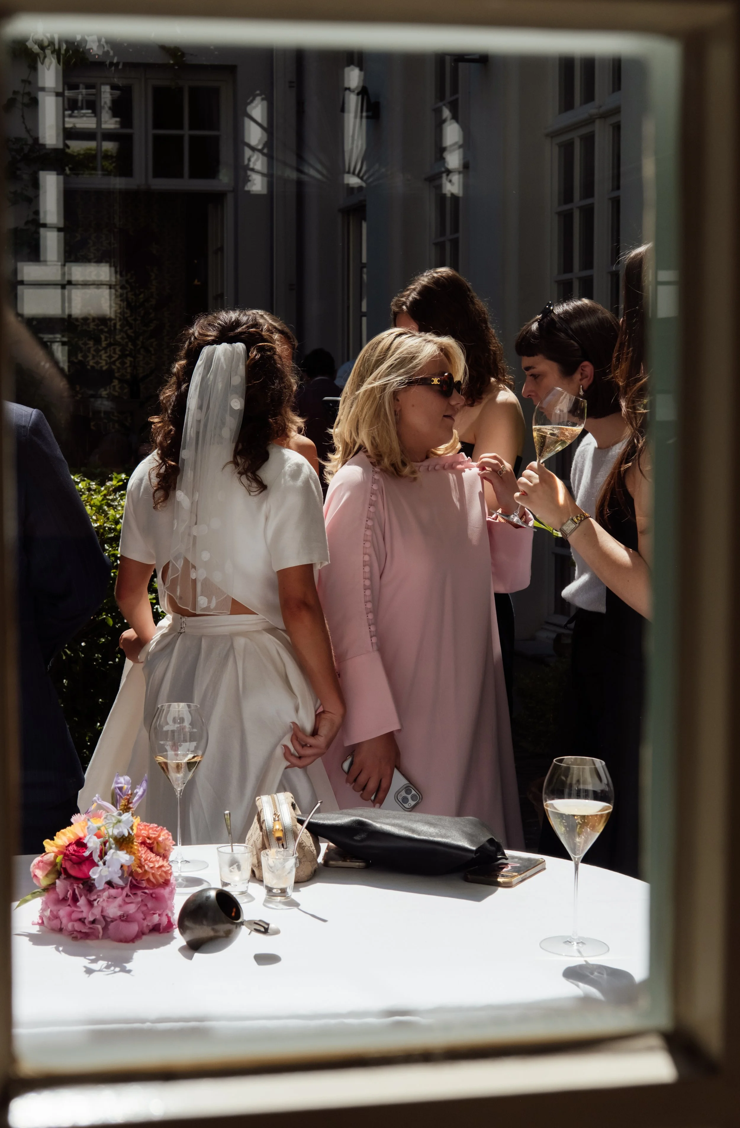 Group of women at an outdoor celebration, one in a white dress with a veil, another in a pink dress with sunglasses, and others chatting, with glasses of champagne on the table.