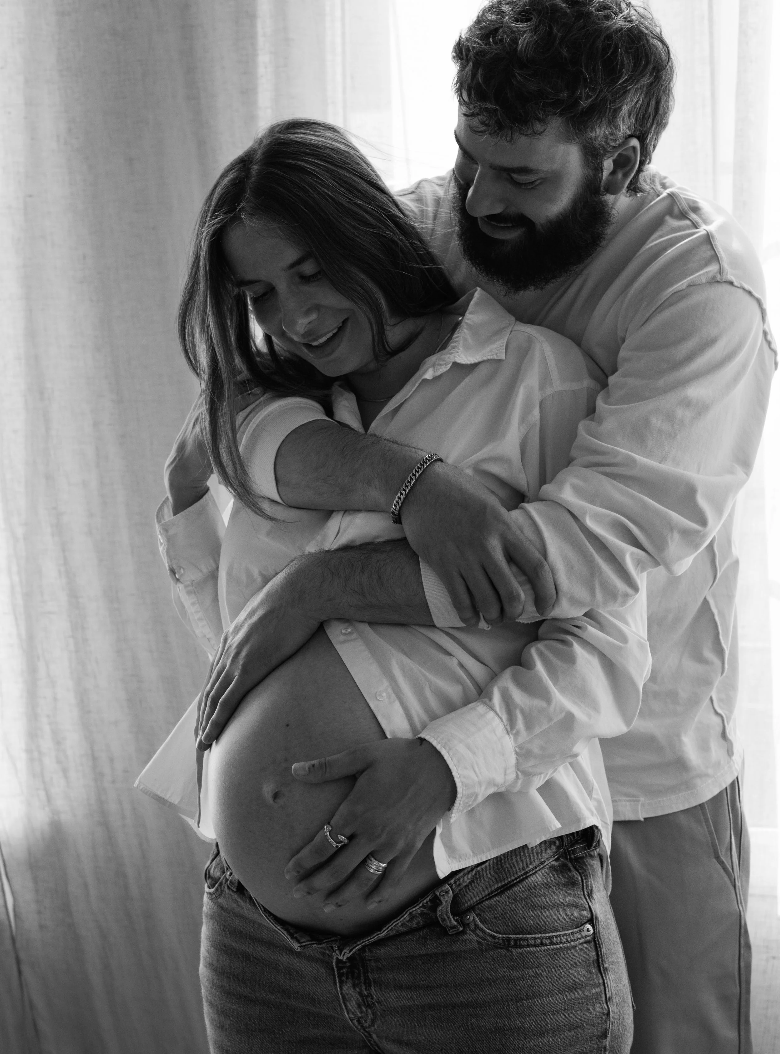 A couple sharing an intimate moment, with the woman pregnant, with the man embracing her from behind, both smiling, near a window.