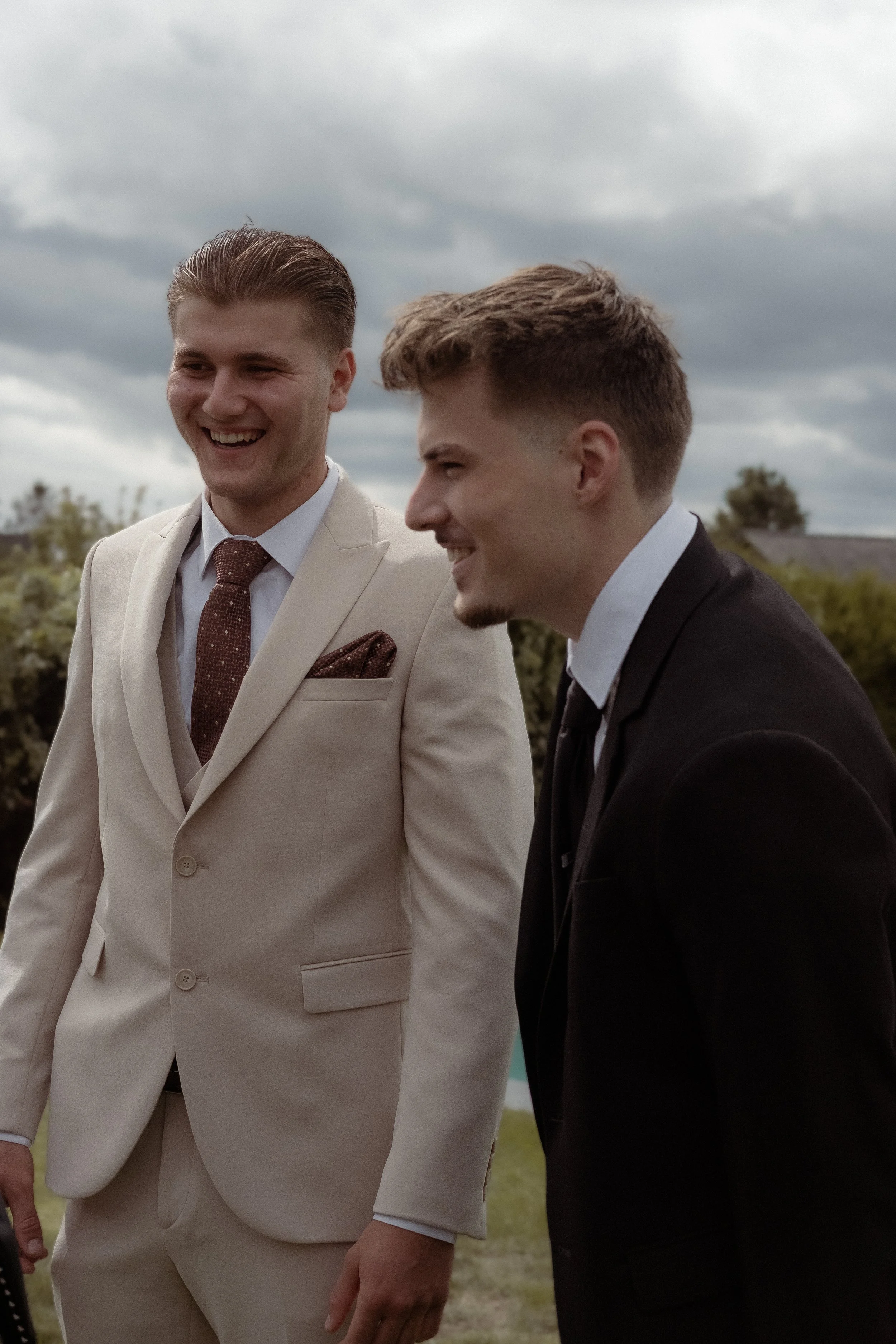 Two young men dressed formally smiling outdoors under cloudy sky, one in a beige suit and the other in a black suit.