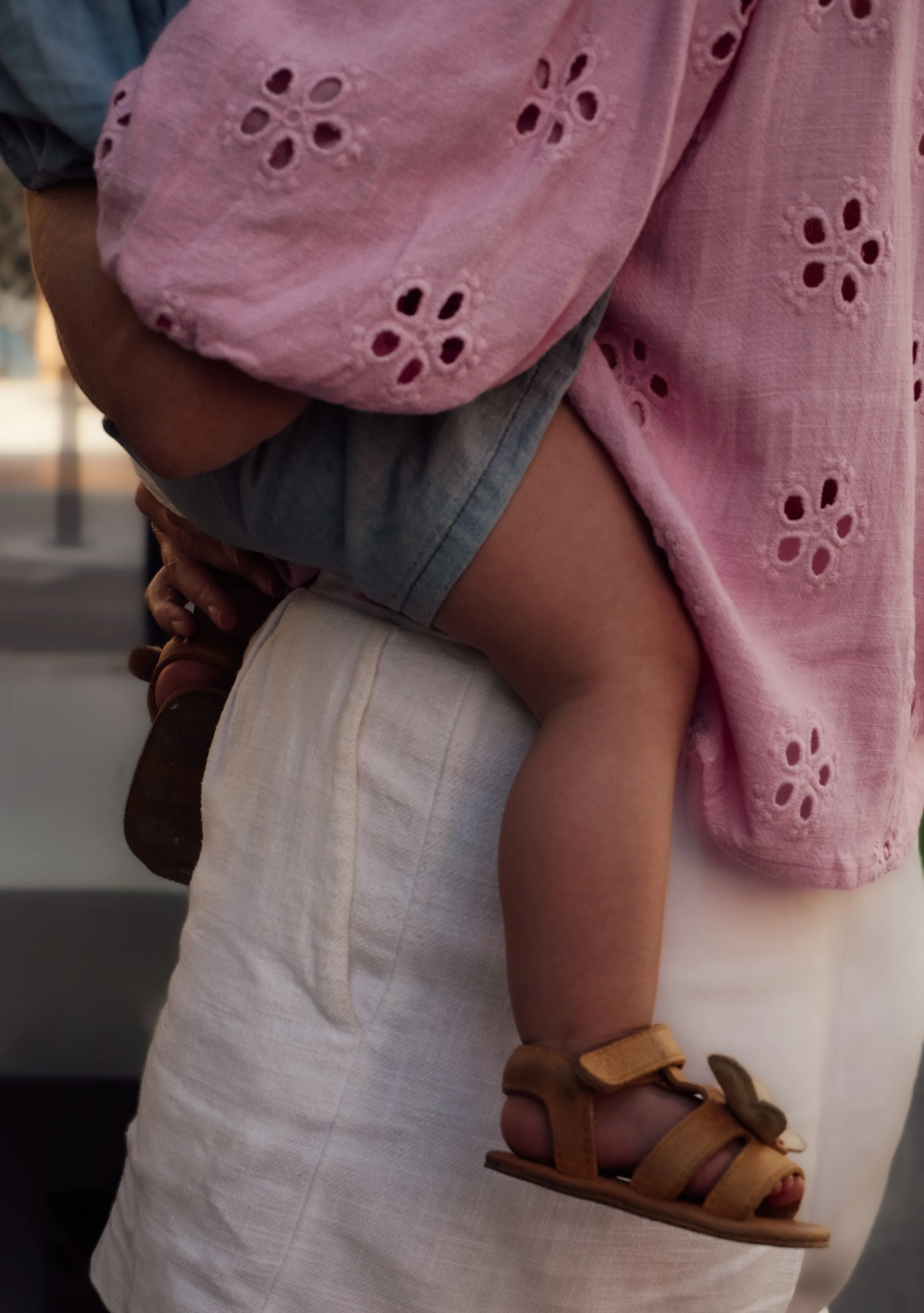 A person wearing a pink eyelet top, beige shorts, and tan sandals is being carried in someone's arms.