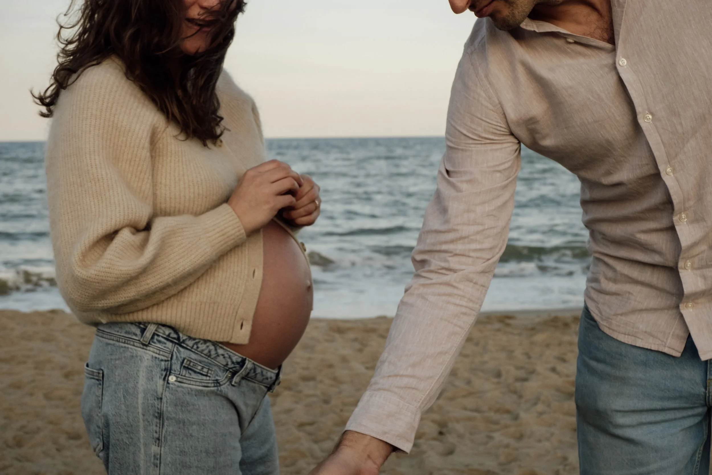 A pregnant woman and a man holding hands on the beach, with the woman looking at her belly.