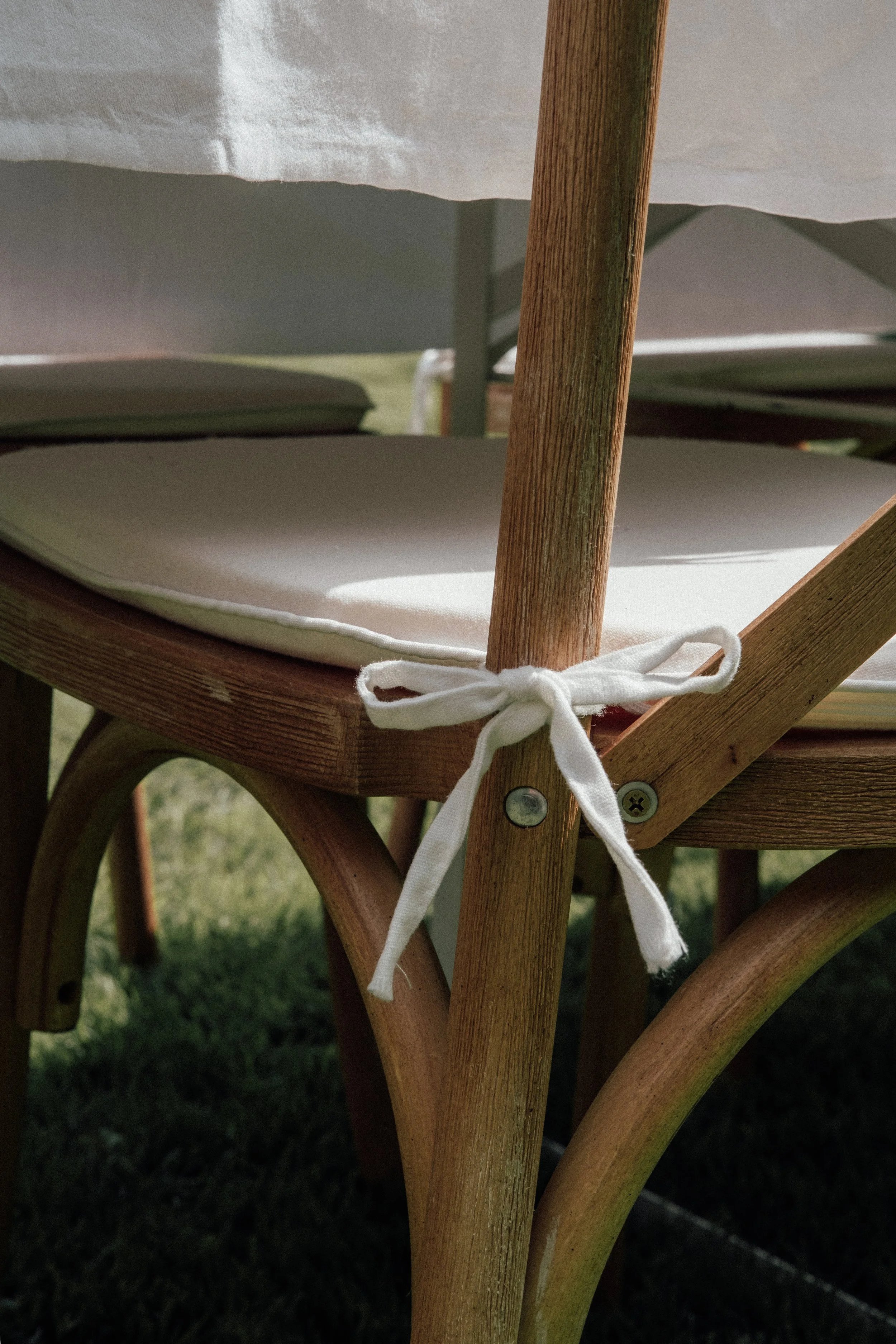 Close-up of a wooden outdoor chair with a white cushion tied to the seat and a white tablecloth draped over a table in the background, set on grass.
