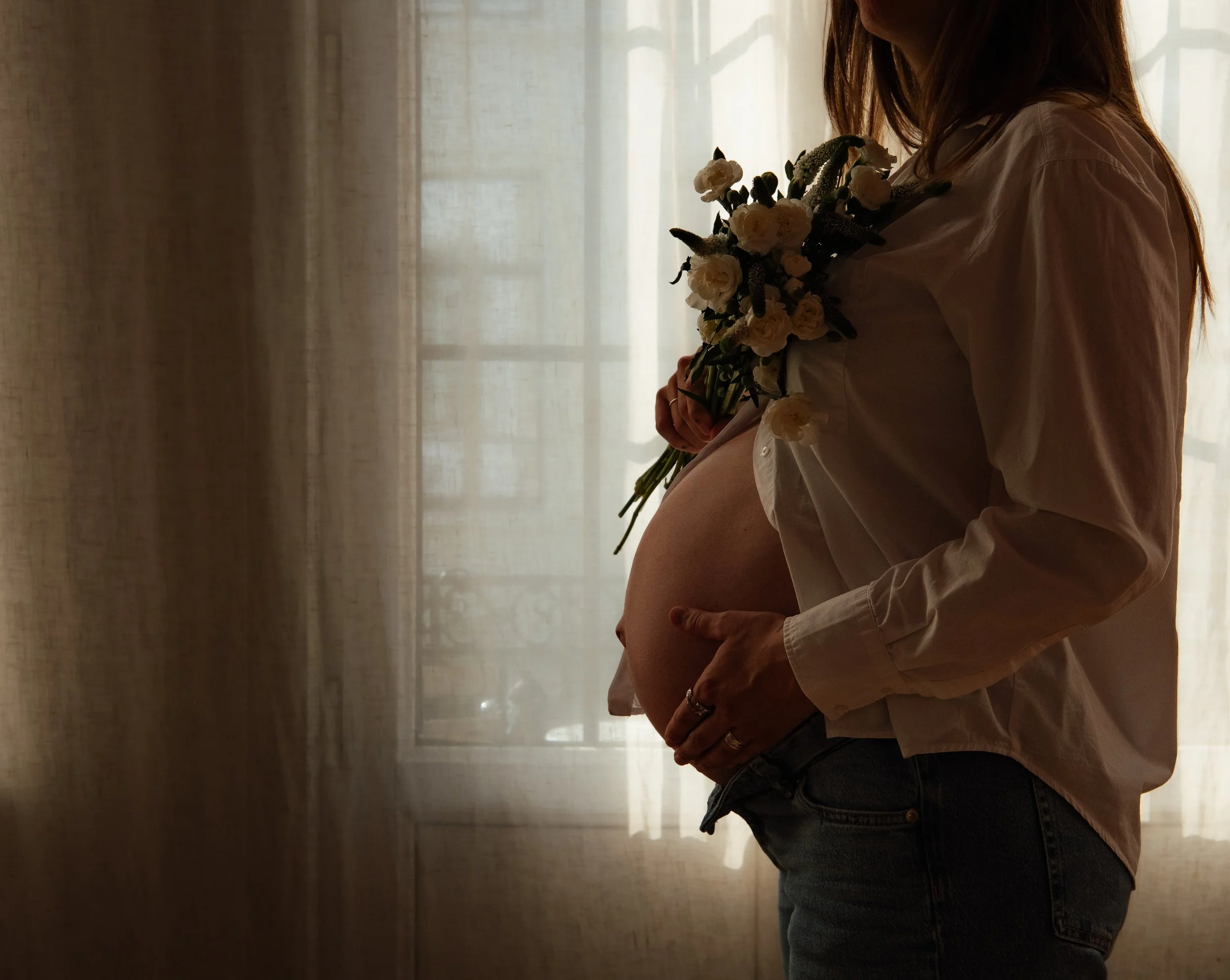 A woman holding a bouquet of flowers, standing by a window with sheer curtains, backlit creating a silhouette effect.