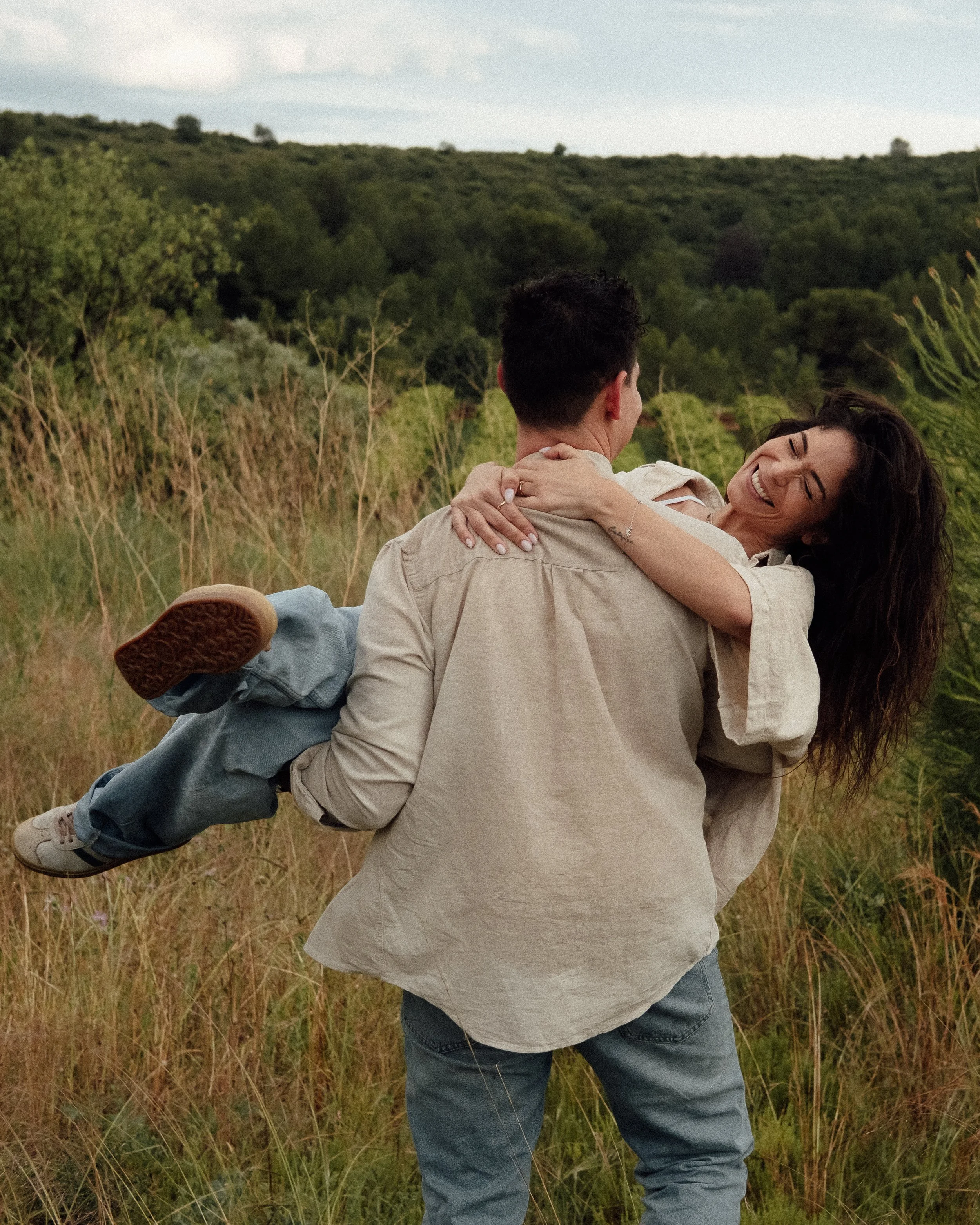 A couple smiling and embracing outdoors in a grassy field with trees in the background, one person being carried by the other.