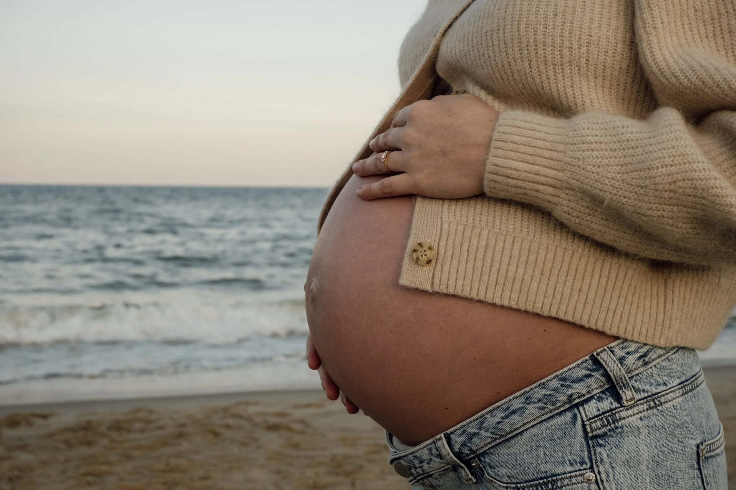 Close-up of a pregnant woman on the beach, wearing a beige sweater and denim shorts, holding her belly with one hand, with the ocean in the background.